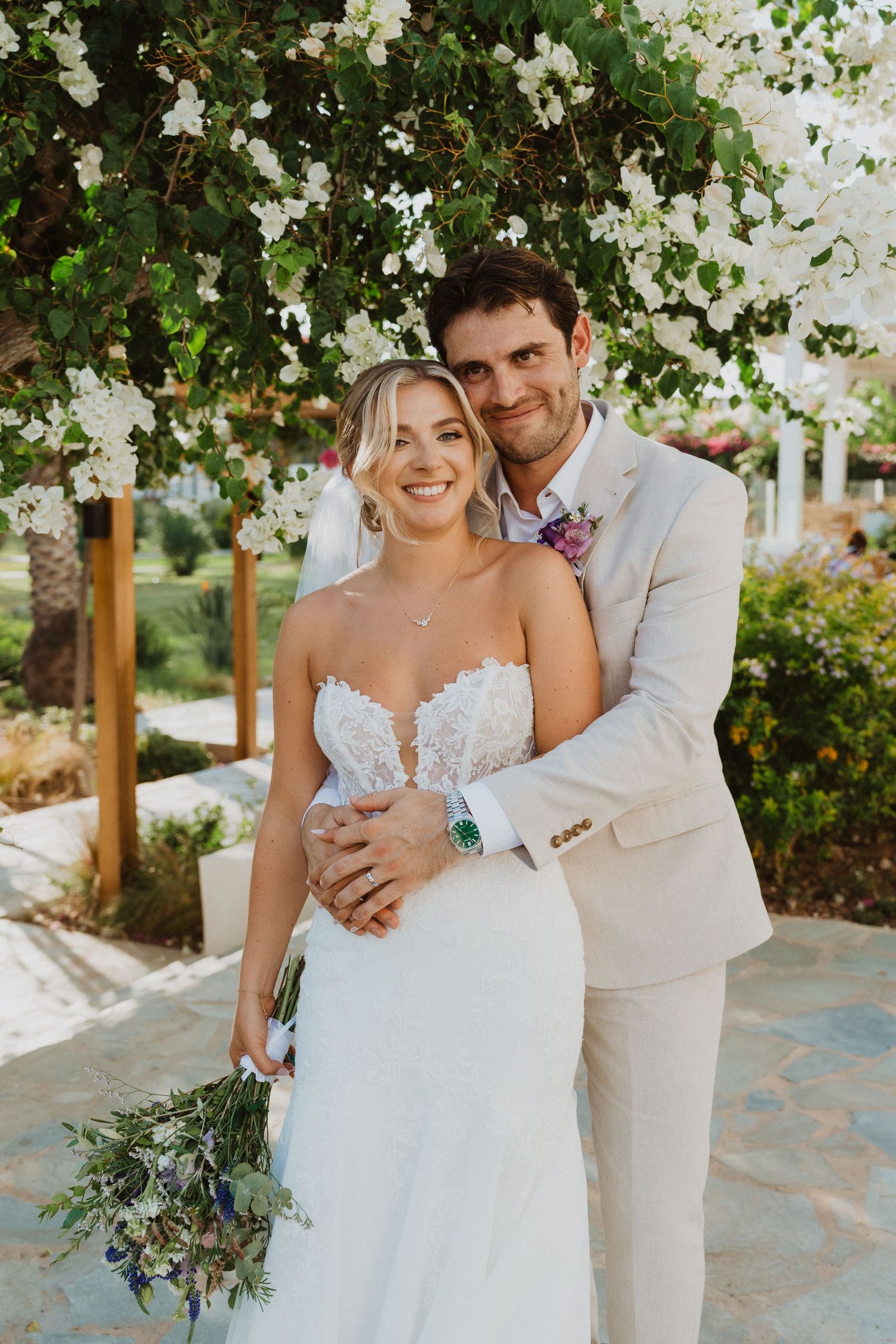 Wedding couple poses under flowering trees on stone pathway in garden setting.