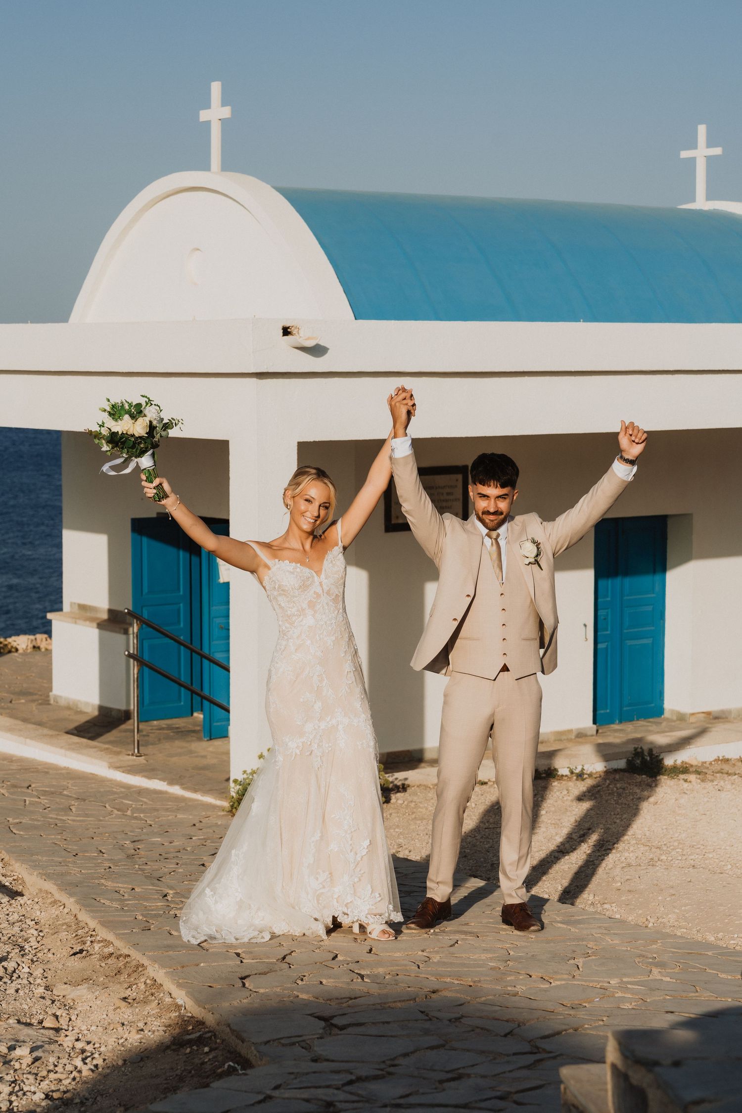 Newlyweds celebrate with raised arms in front of a white Mediterranean church overlooking the ocean.