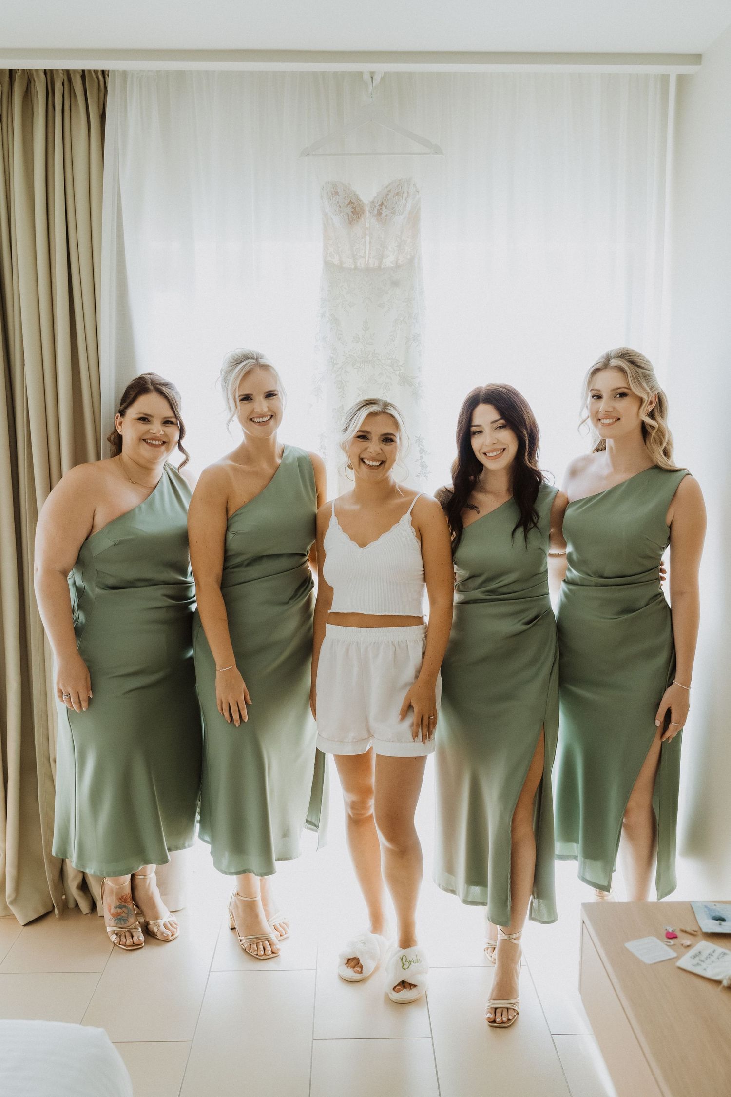 Group wearing sage green bridesmaid dresses pose with someone in white loungewear.