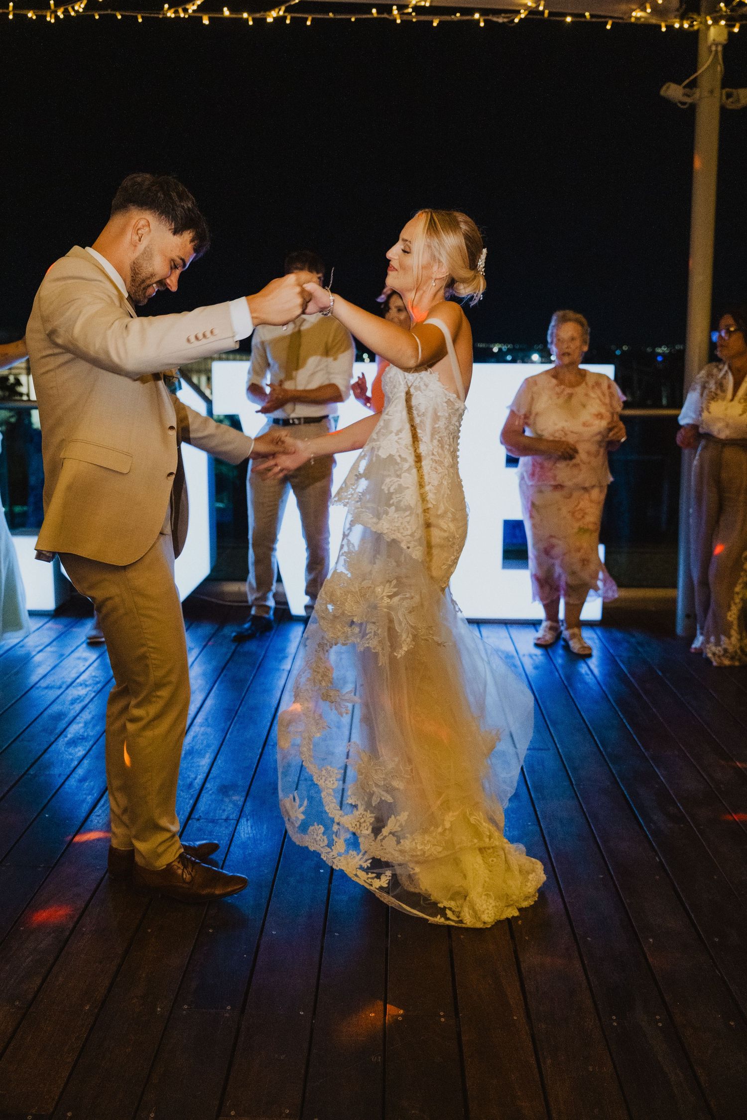 Couple dancing on wooden deck during evening wedding reception with string lights overhead.