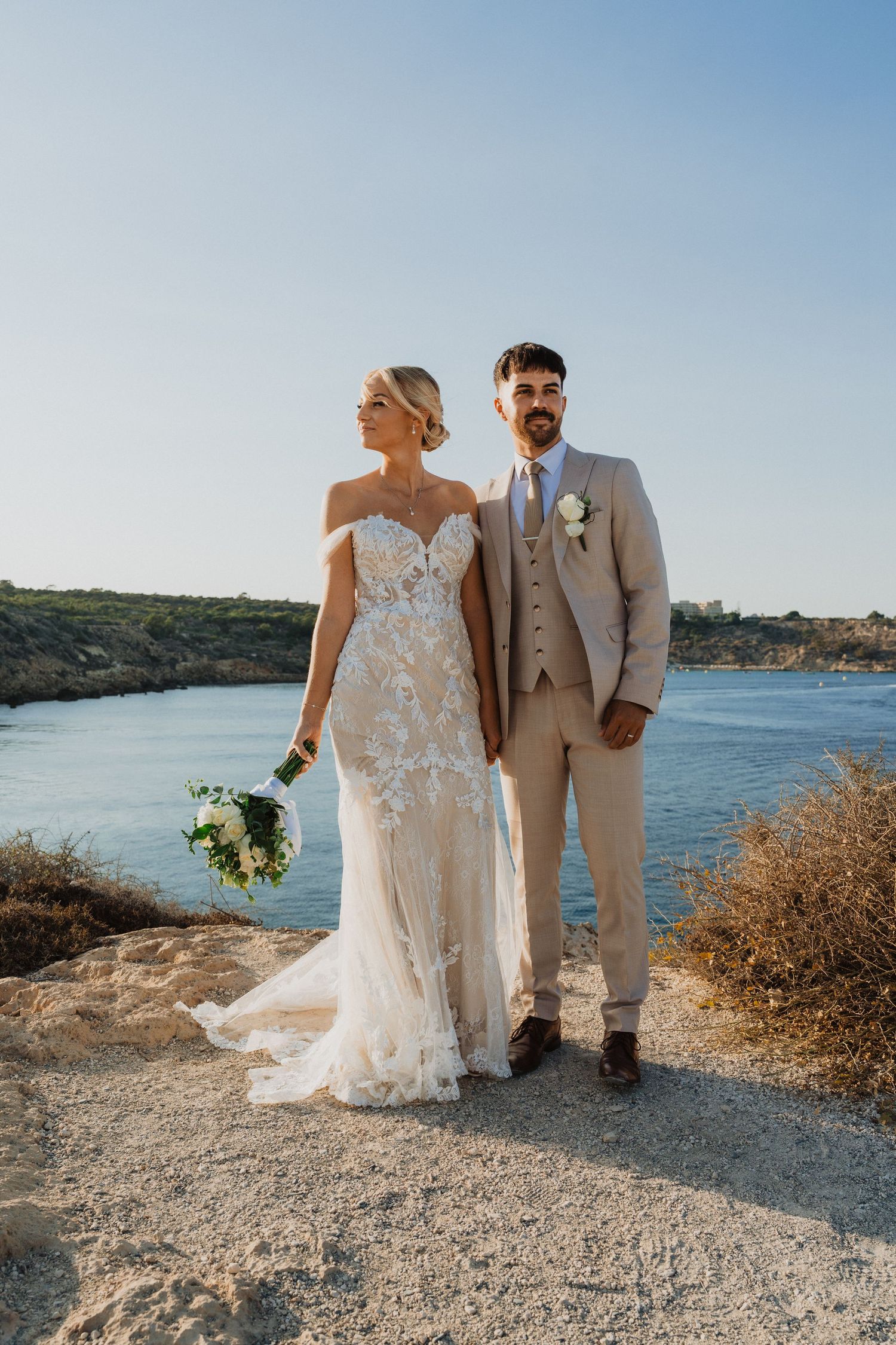 A couple poses for wedding photos on a rocky coastal path overlooking a serene blue bay.