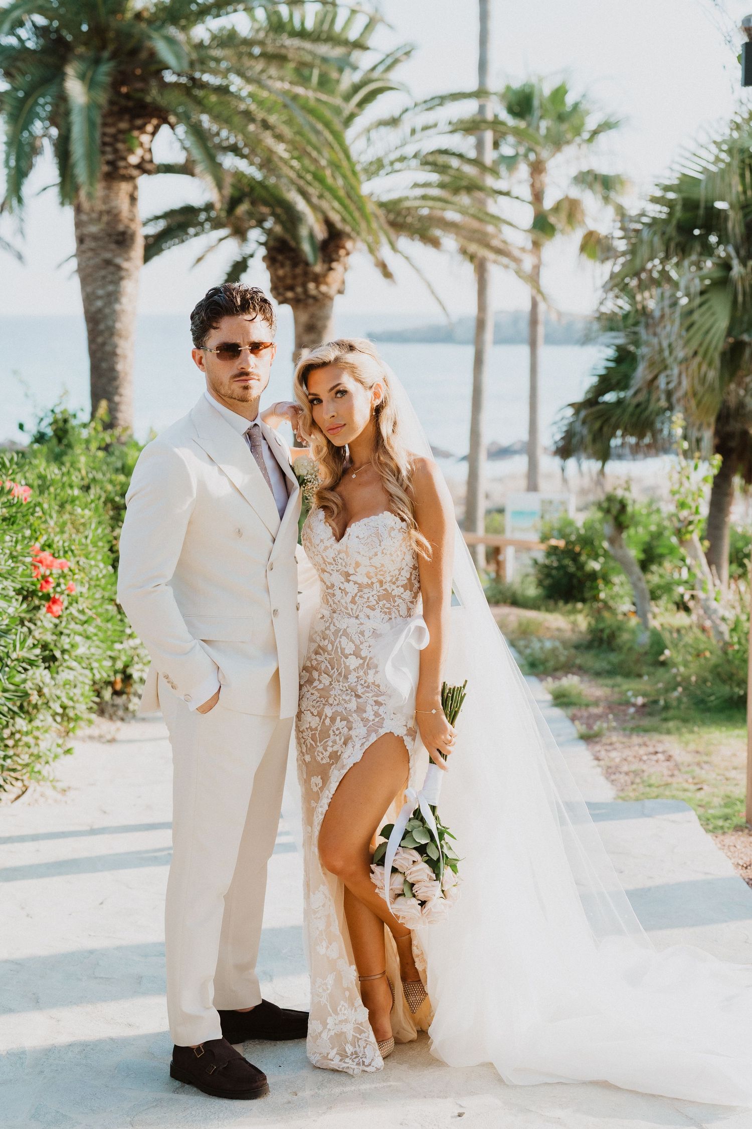 A couple poses together in elegant white wedding attire on a palm-lined beach walkway.