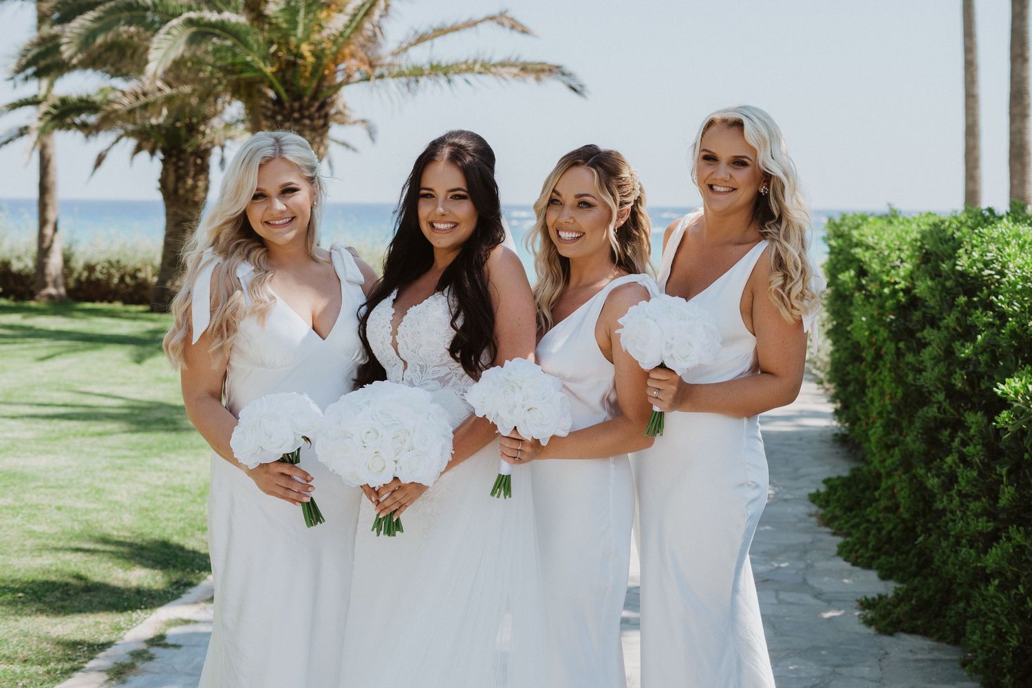 Wedding party in white dresses walking together along a palm tree-lined path in a tropical setting.