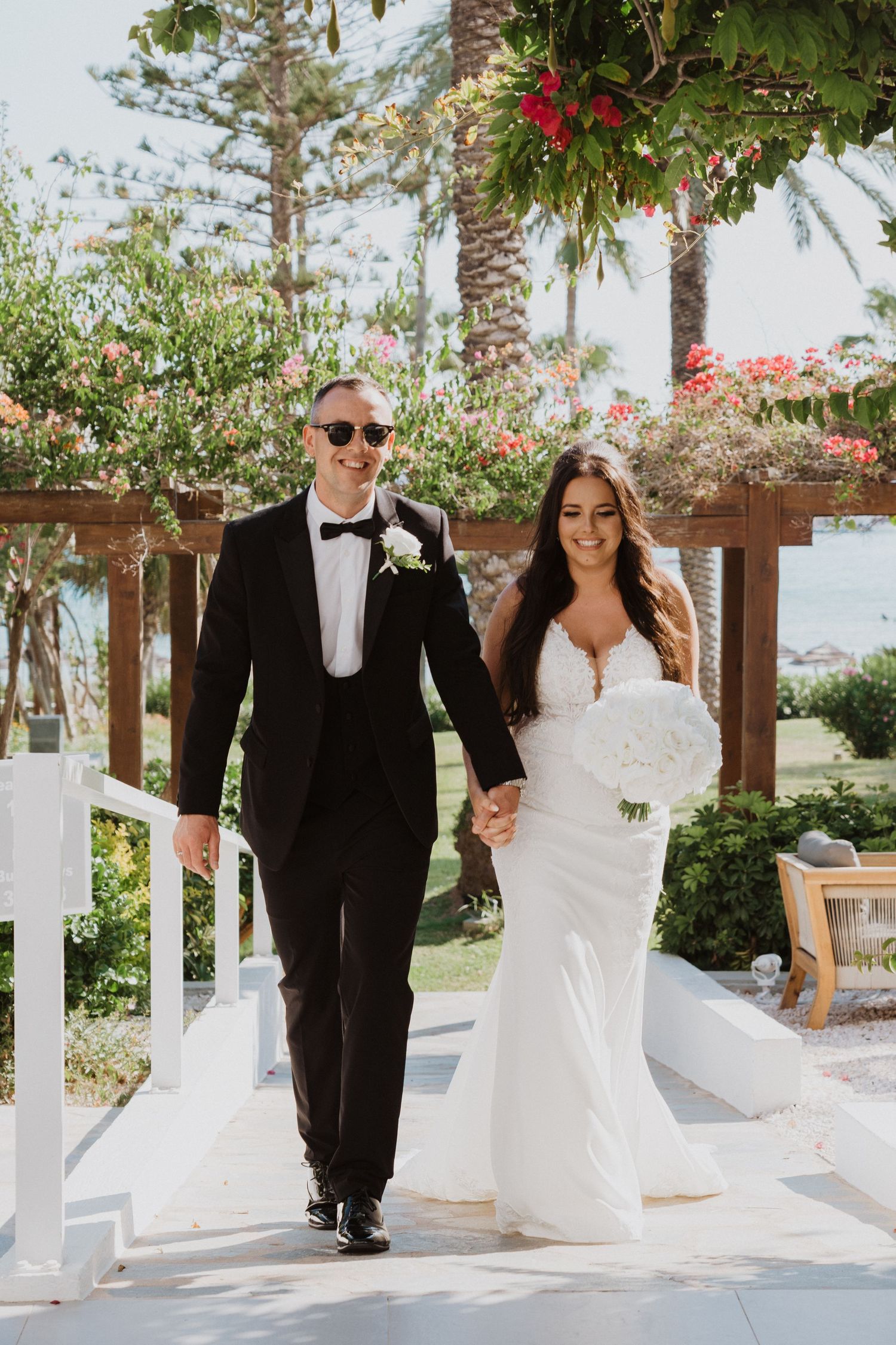 A couple celebrates their wedding day under a white pergola surrounded by tropical landscaping.