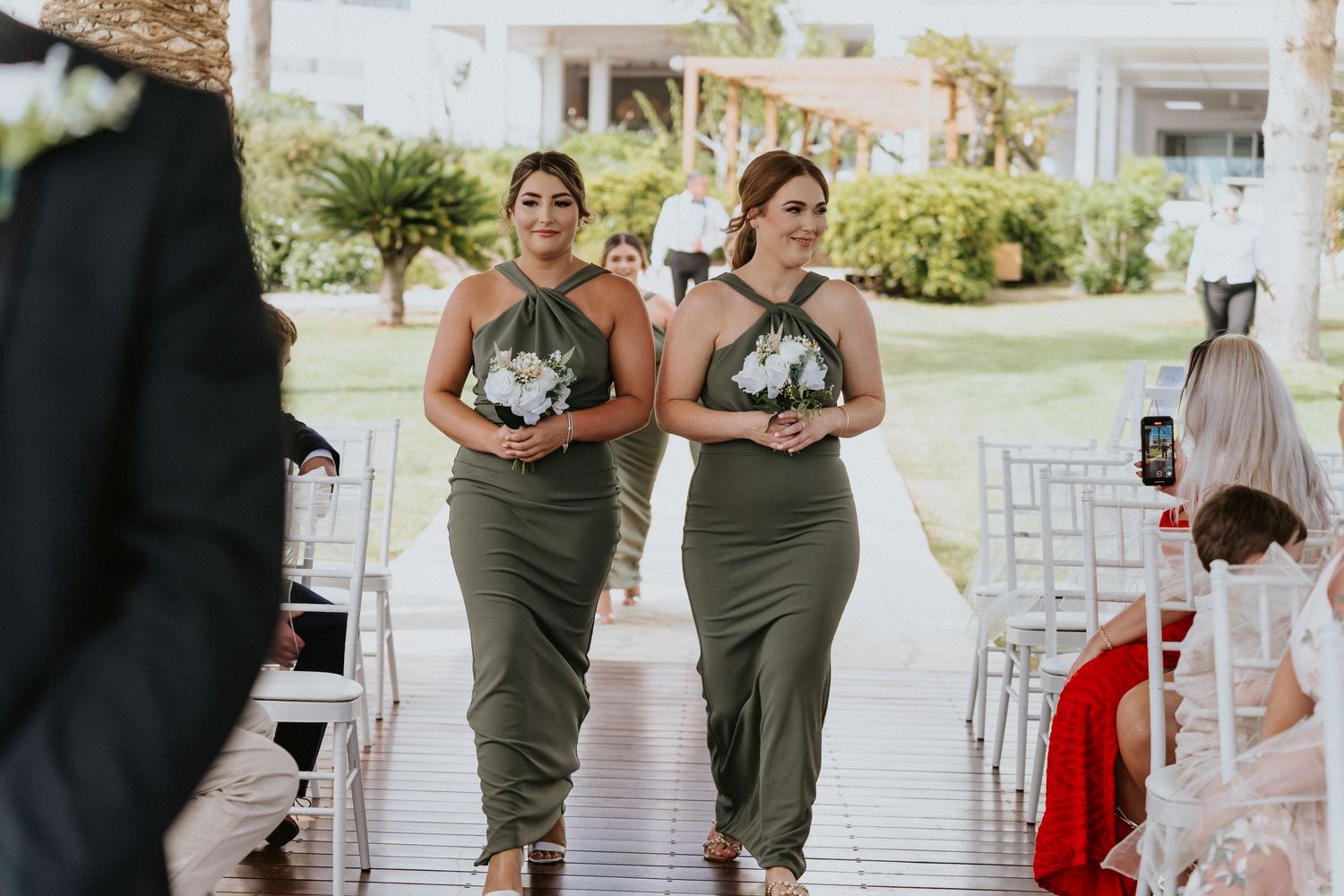 Wedding party members walking down the aisle in olive green dresses carrying white bouquets.