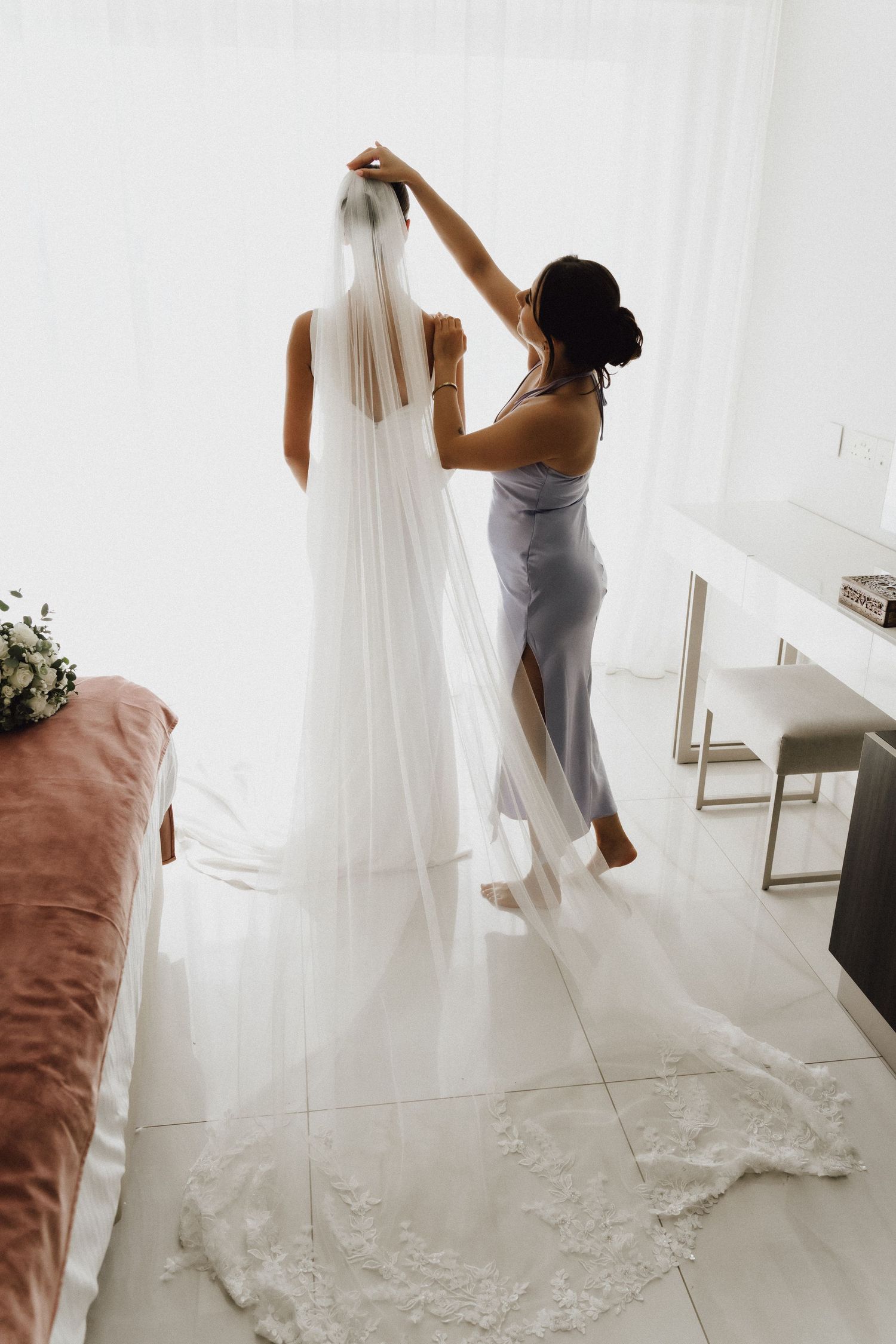 A person in gray helps adjust a long flowing white wedding veil in a bright modern room.