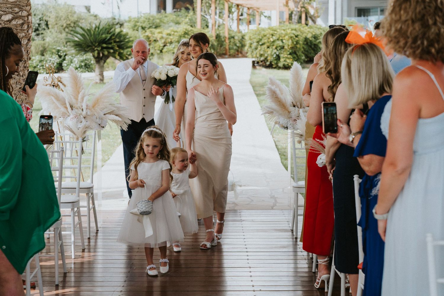 Bridal party processional on covered wooden deck with guests watching from both sides.