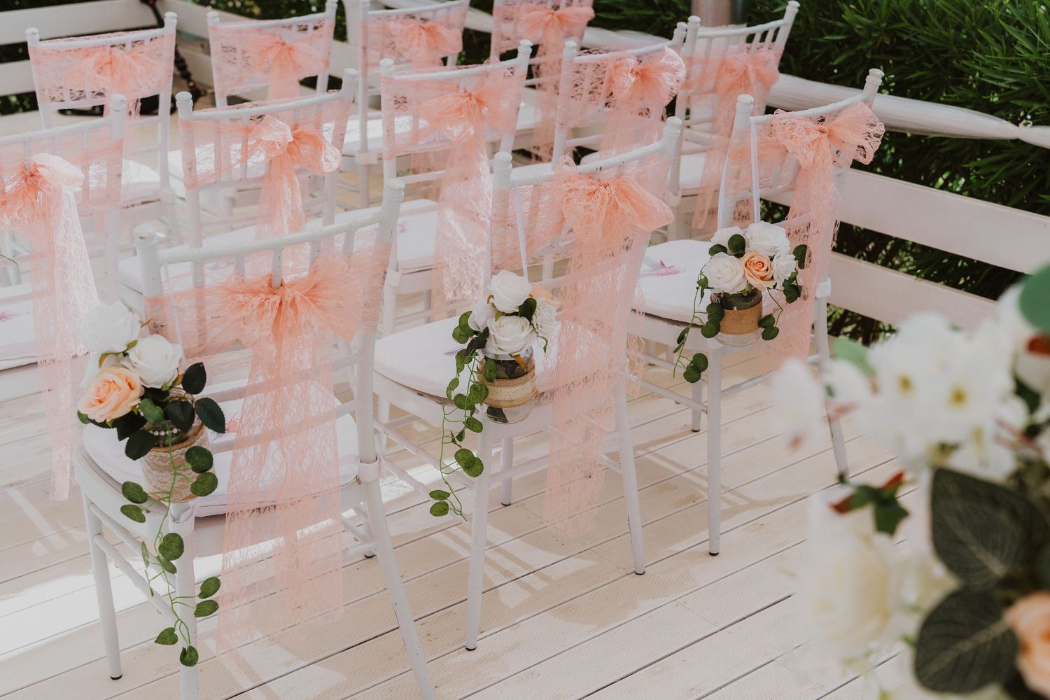 Clear acrylic chairs decorated with delicate pink ribbons and floral arrangements for an elegant outdoor wedding ceremony.