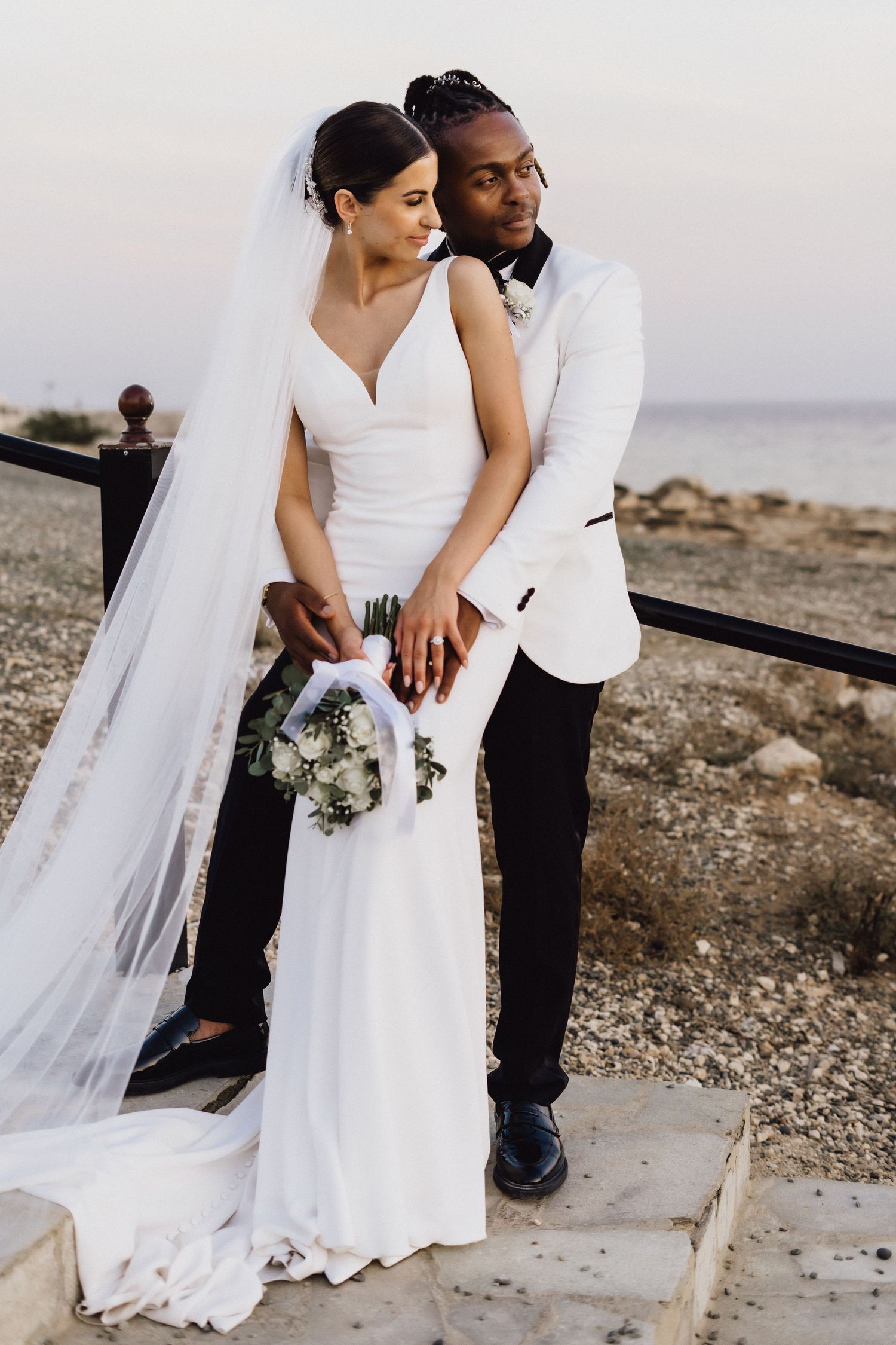 Elegant wedding couple in white and black attire pose against Mediterranean sunset.