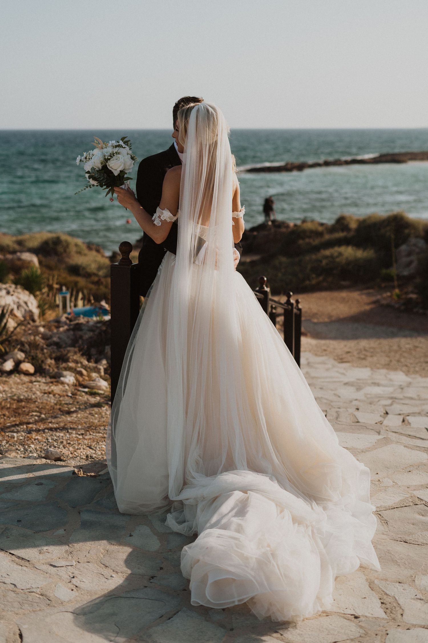 Bride in flowing white gown holds bouquet overlooking coastal Mediterranean landscape.