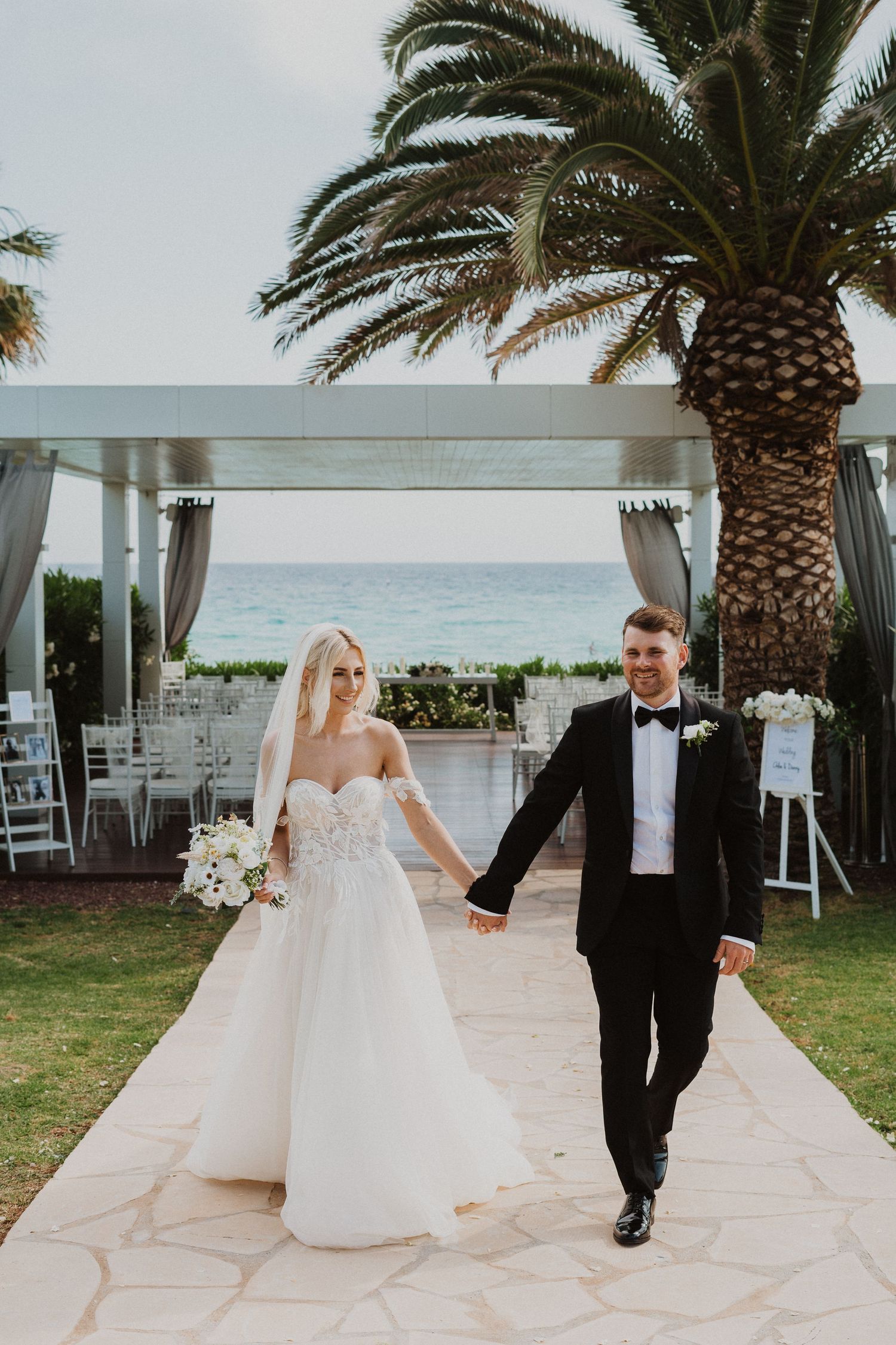 Couple holds hands while walking down a stone path at their beachside wedding venue.