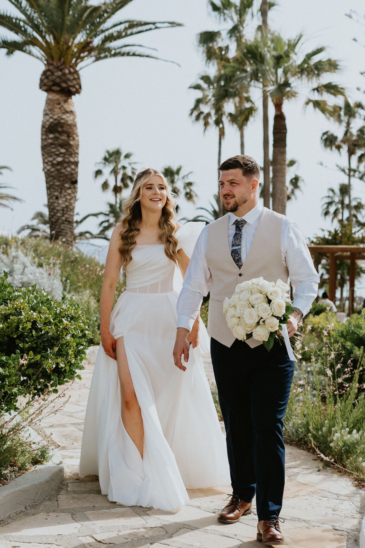 Wedding couple walks down palm-lined path with bride in flowing white dress carrying bouquet.