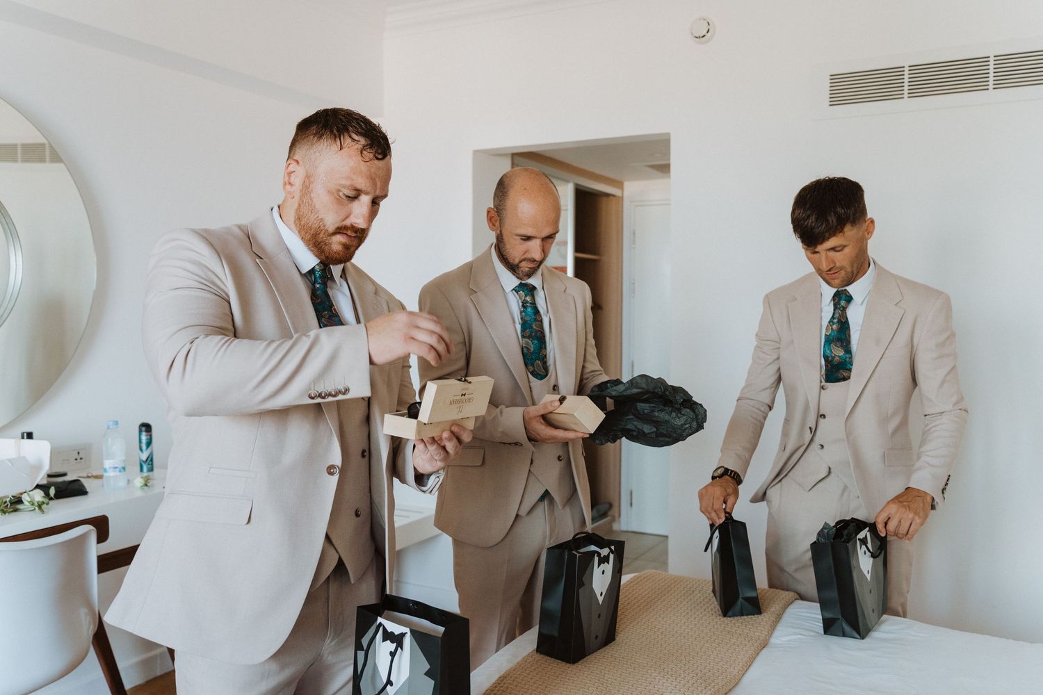Three men in matching beige suits and teal ties getting ready together in a bright room.