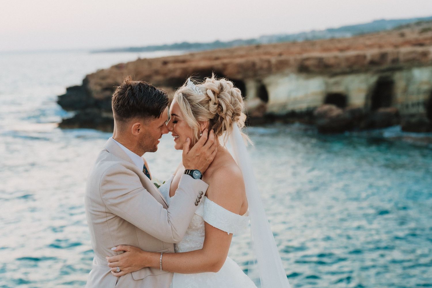 A couple shares a romantic moment by turquoise ocean waters near coastal rock formations at sunset.