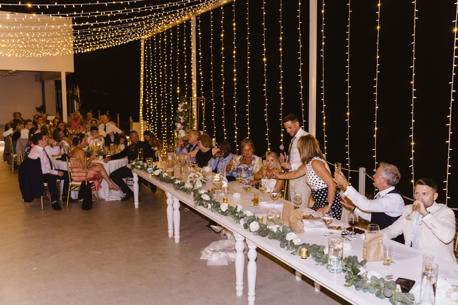 Long reception tables decorated with greenery and string lights overhead at wedding celebration.