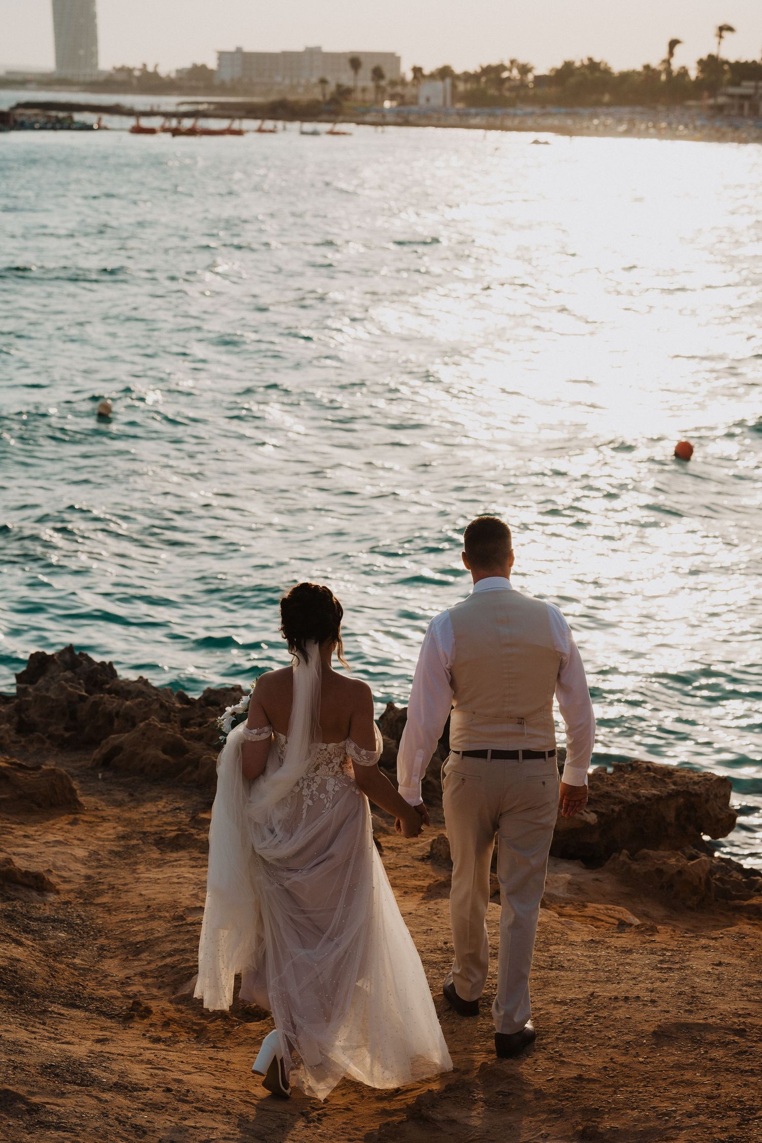 A couple stands together at sunset on a rocky beach overlooking turquoise ocean waters and distant buildings.