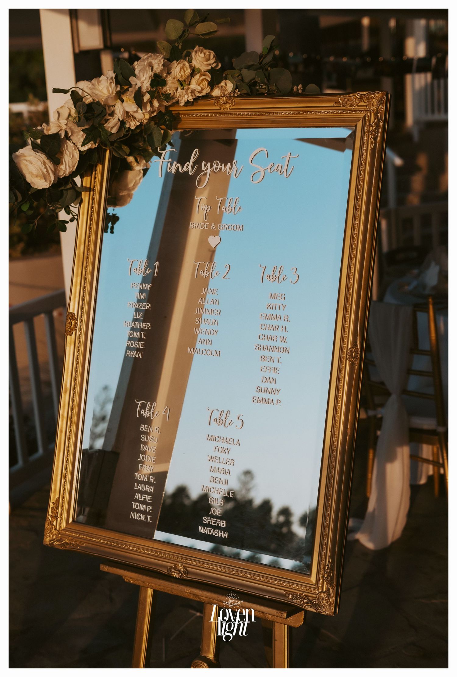 Gold framed mirror seating chart decorated with white roses on wooden easel.
