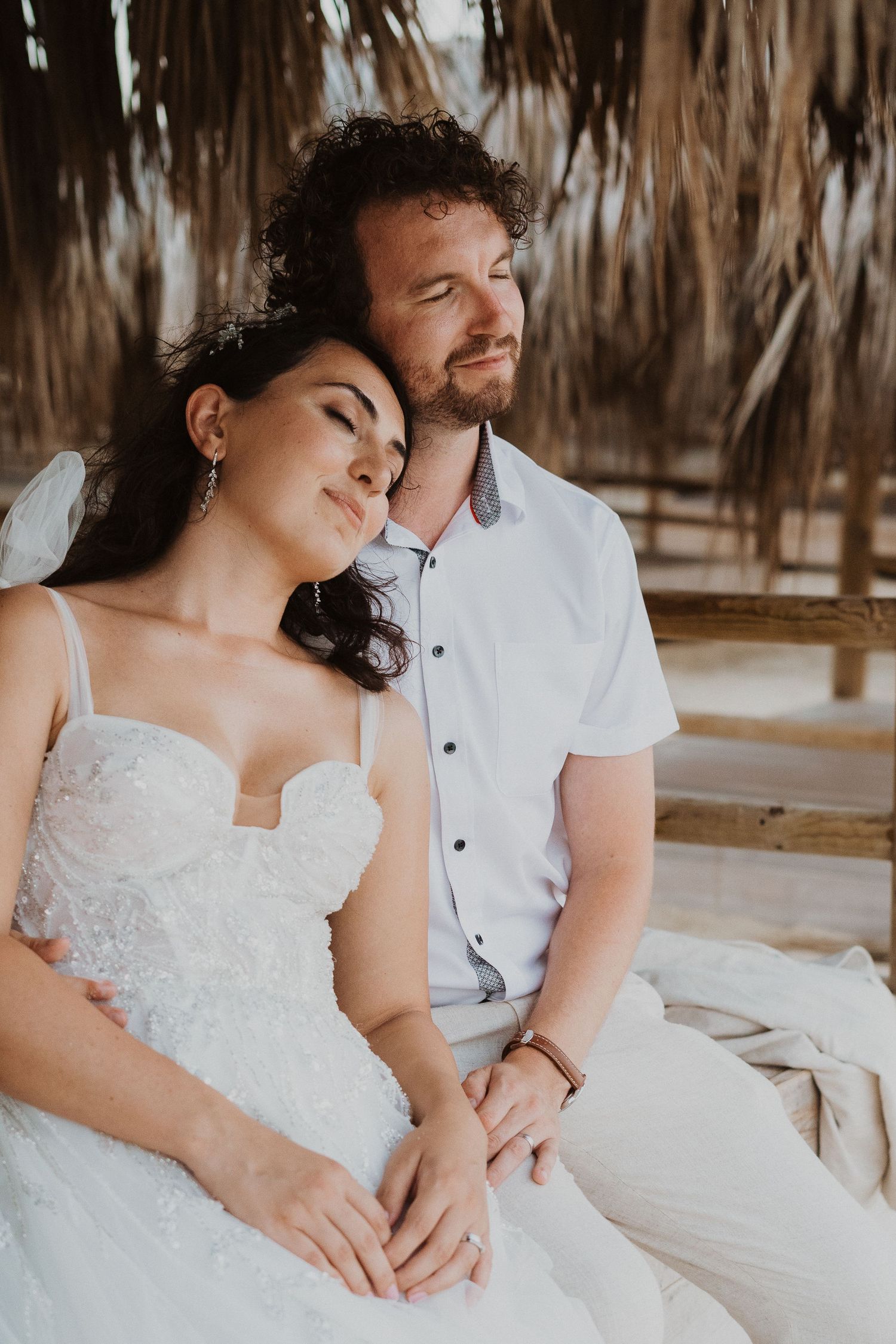A romantic wedding portrait of a couple in white attire under a tropical beach palapa.