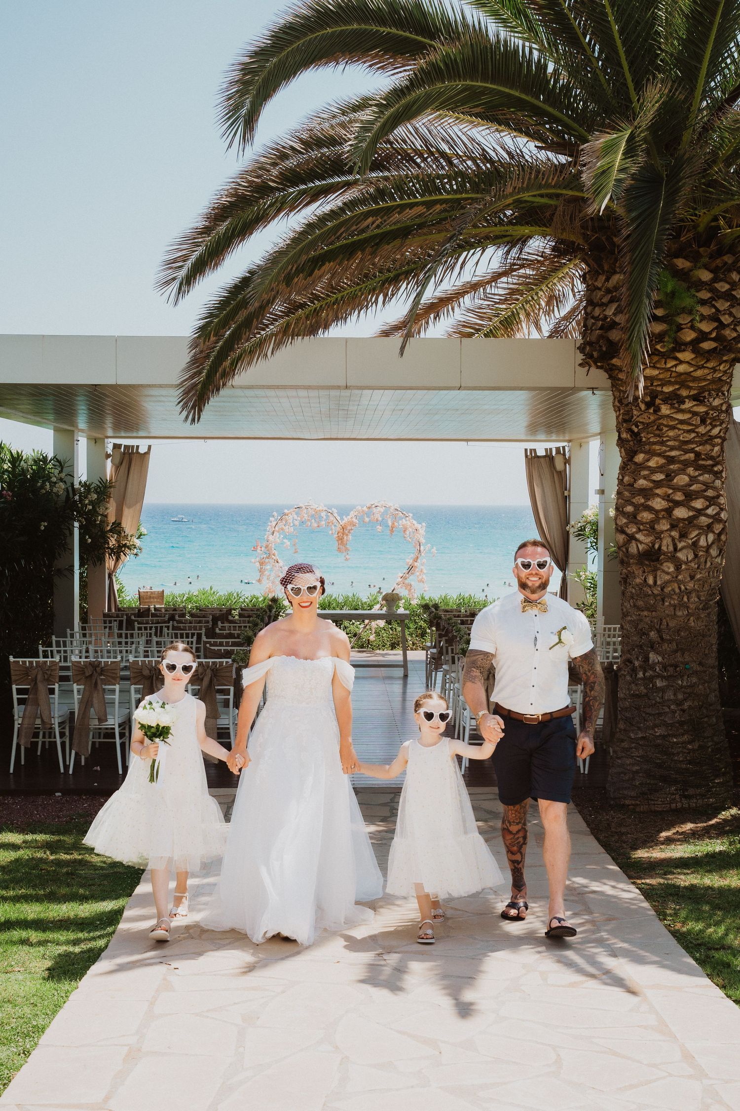Beach wedding scene with family members walking down palm-lined path with heart arch and ocean backdrop.