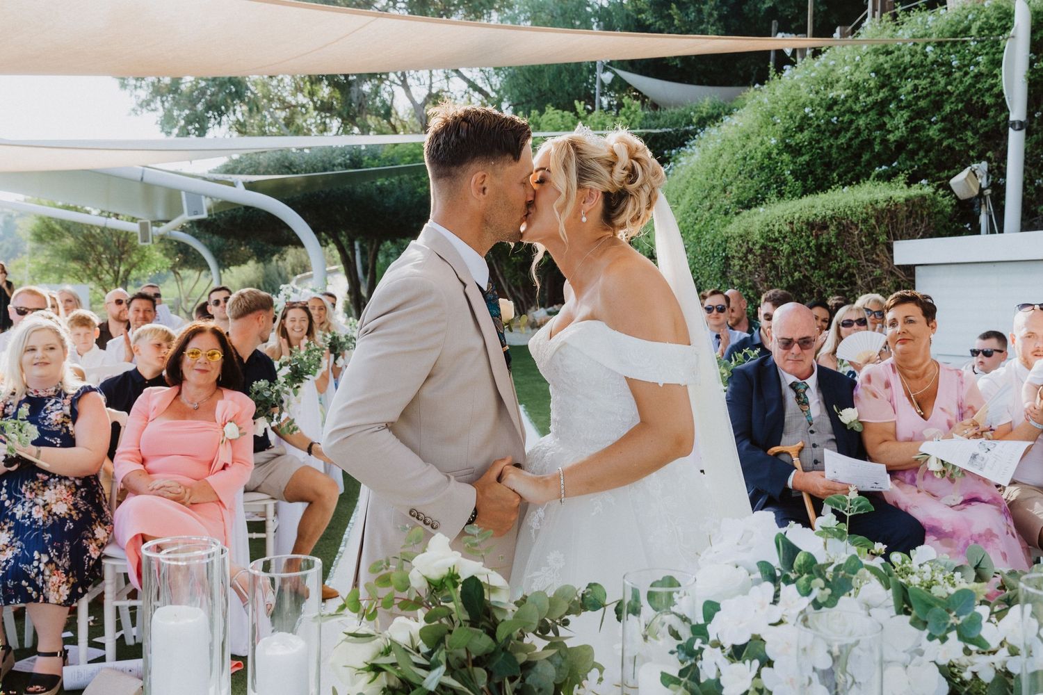 A romantic wedding ceremony moment under outdoor canopies with white floral arrangements and seated guests.