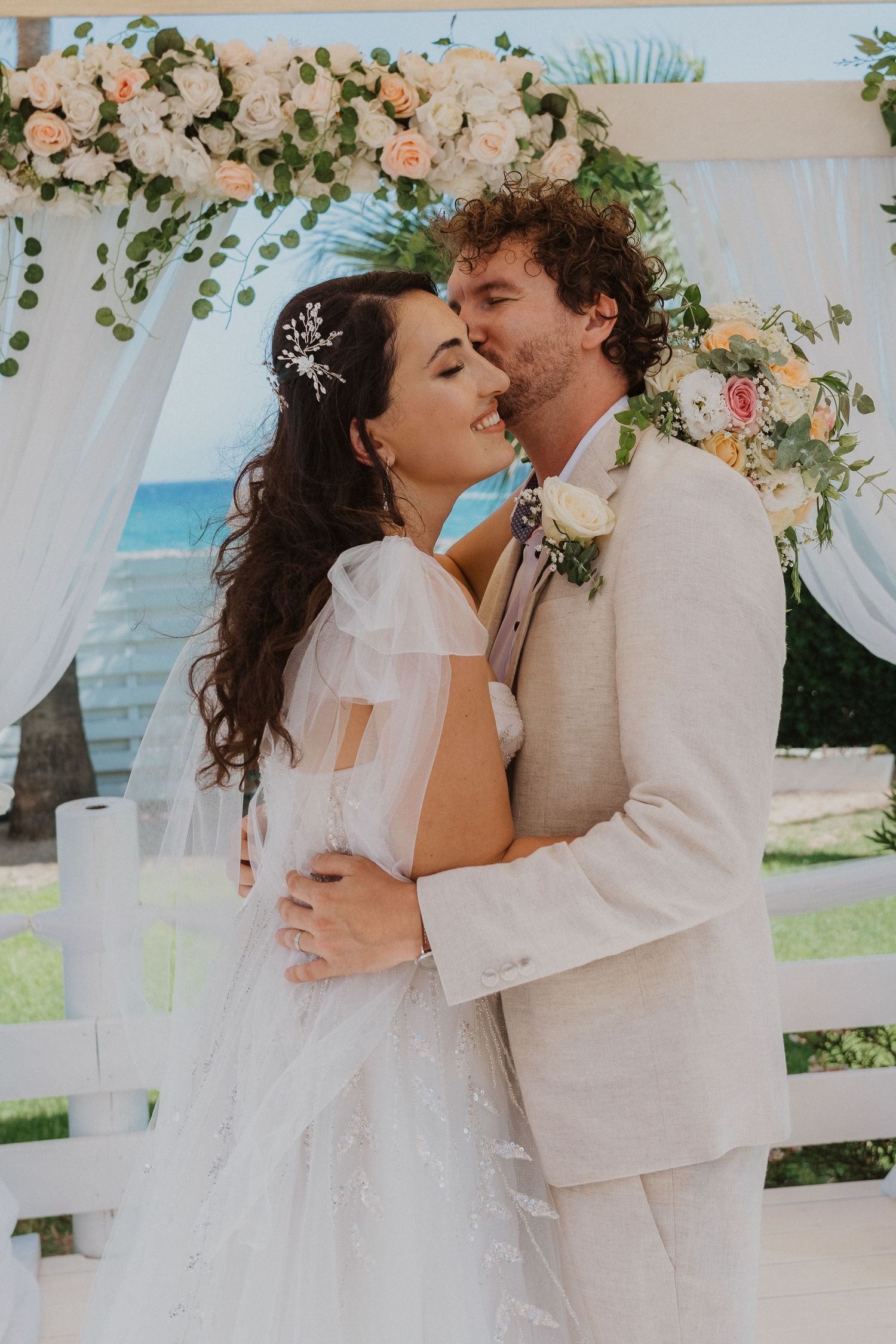 Intimate moment between newlyweds under floral decorated arch with ocean backdrop.