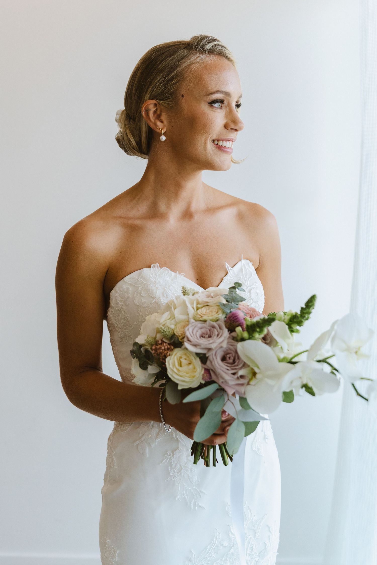 Elegant bride in strapless gown holds romantic bouquet of roses and orchids against white backdrop.