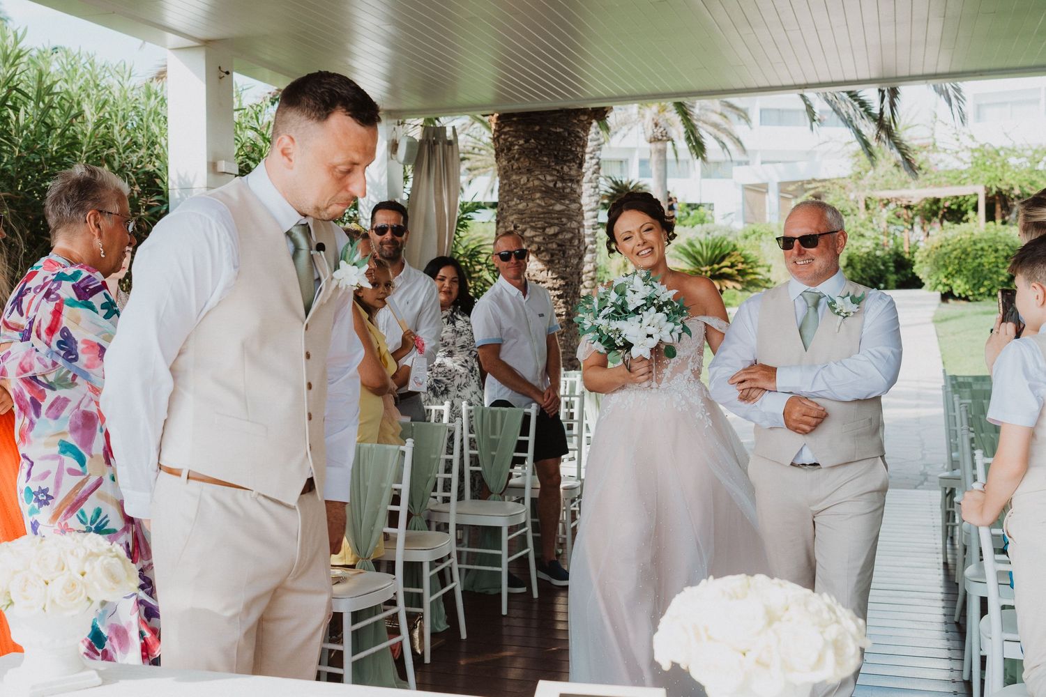 Guests in light summer suits and tropical shirts gather under a covered outdoor patio during a celebration.