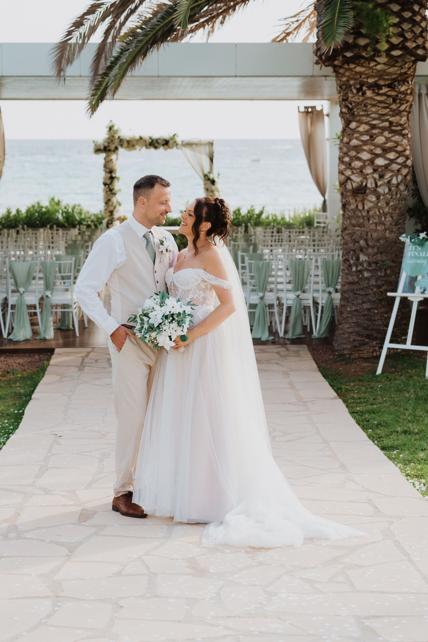 A romantic beachside wedding ceremony setup with palm trees and a white floral arch on a stone pathway.