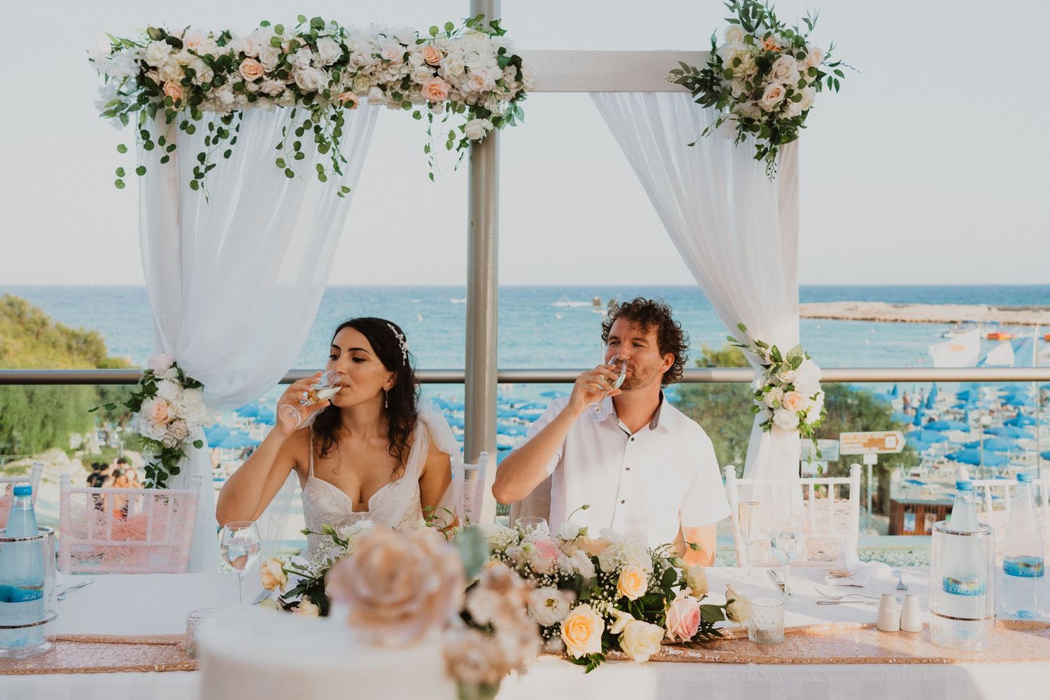 Elegant wedding reception at a beachfront venue with floral arch and white drapes overlooking the ocean.