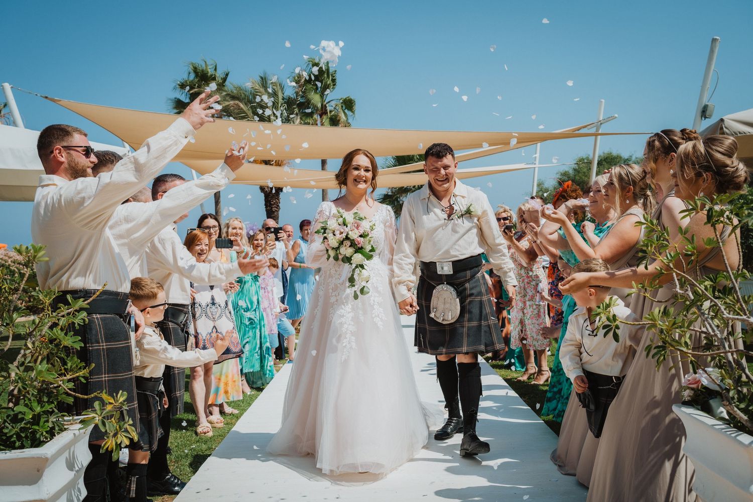 Wedding guests celebrate by throwing confetti at couple walking down aisle at beach ceremony.