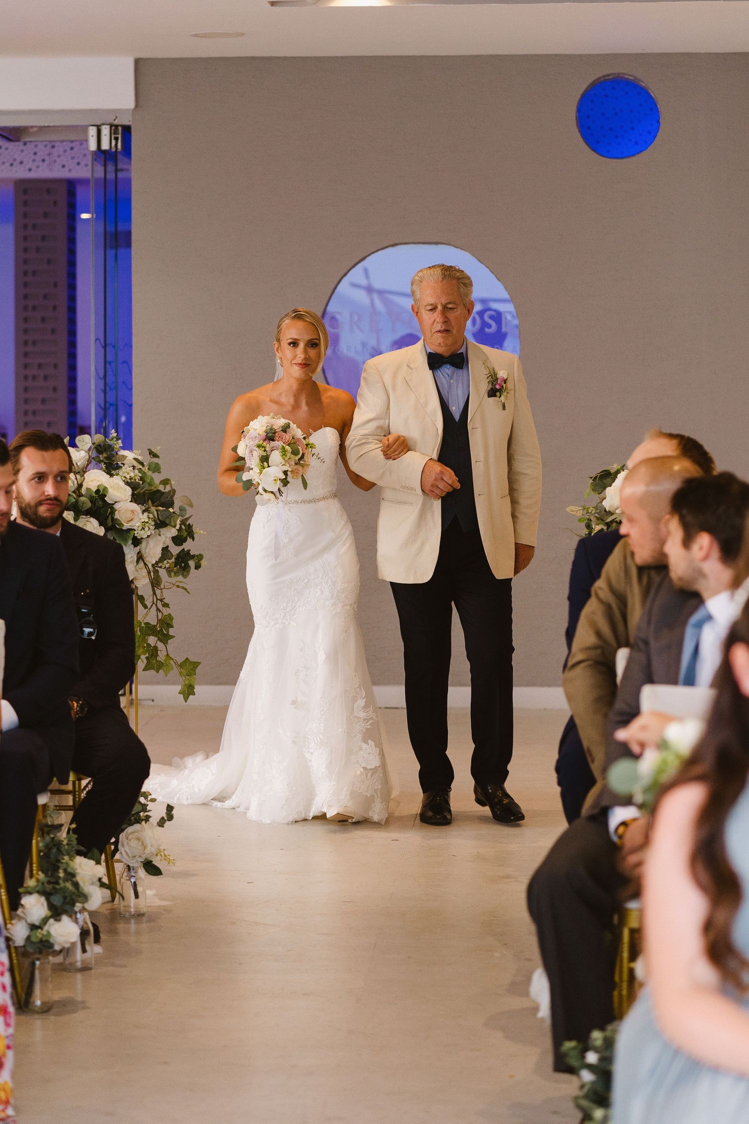 Wedding processional in a modern venue with blue circular window and floral aisle decorations.