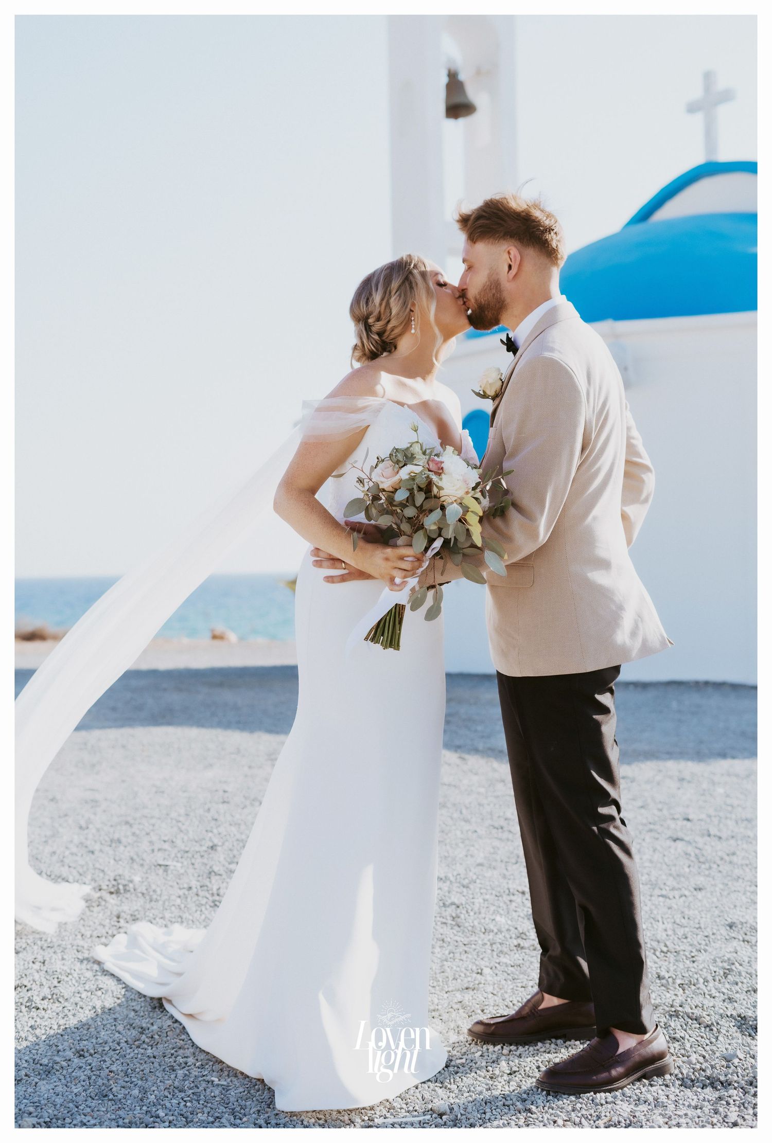 Romantic wedding kiss captured in front of traditional Greek church with blue dome.