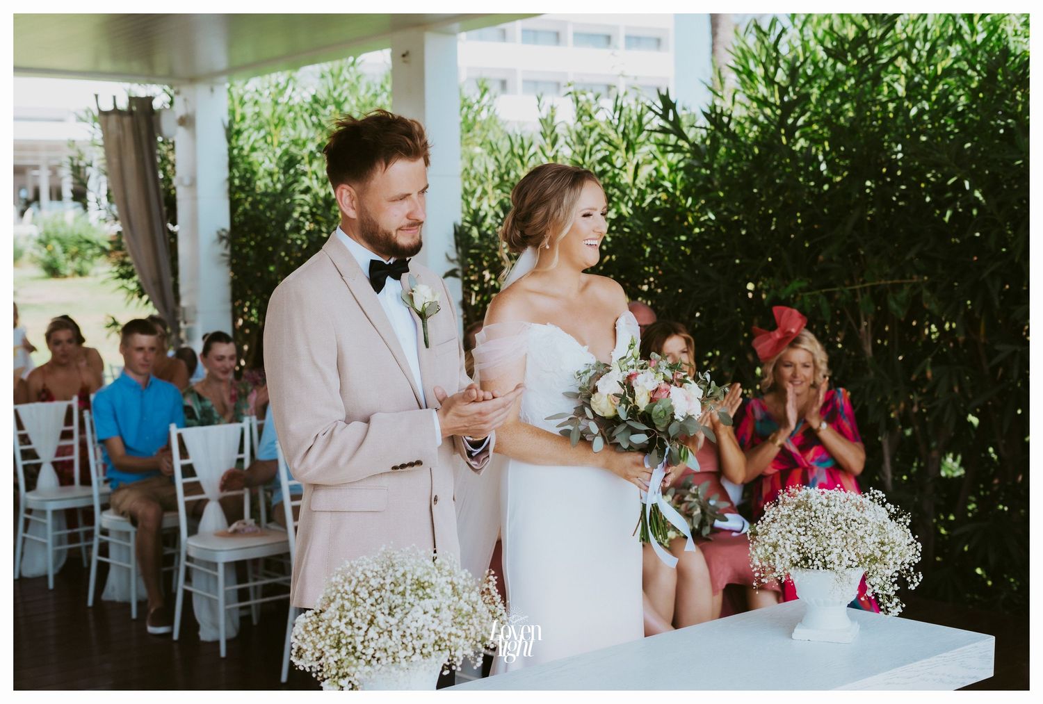 A couple stands at a wedding ceremony table decorated with baby's breath flowers under an outdoor pavilion.