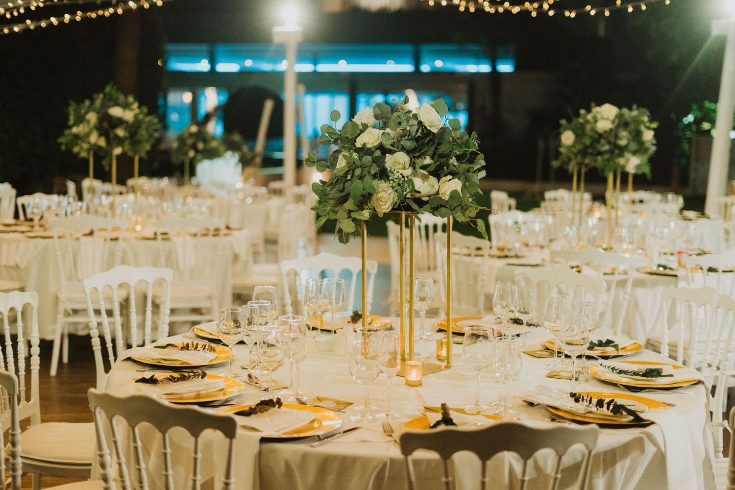 Elegant wedding reception setup with round tables, white chairs, and tall centerpiece floral arrangements at night.