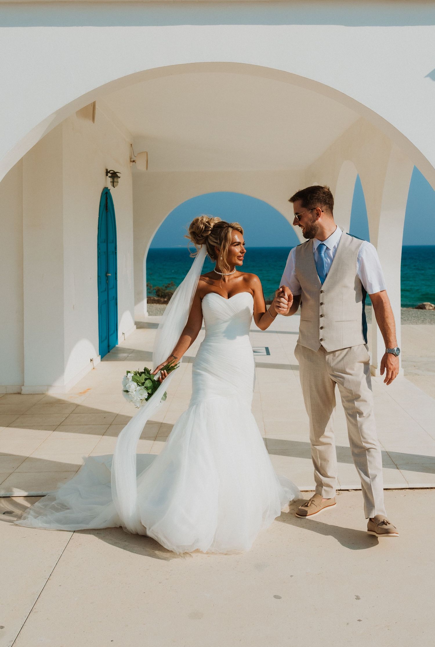 Bride and groom walking hand in hand at a Mediterranean seaside venue with white arches and blue door.