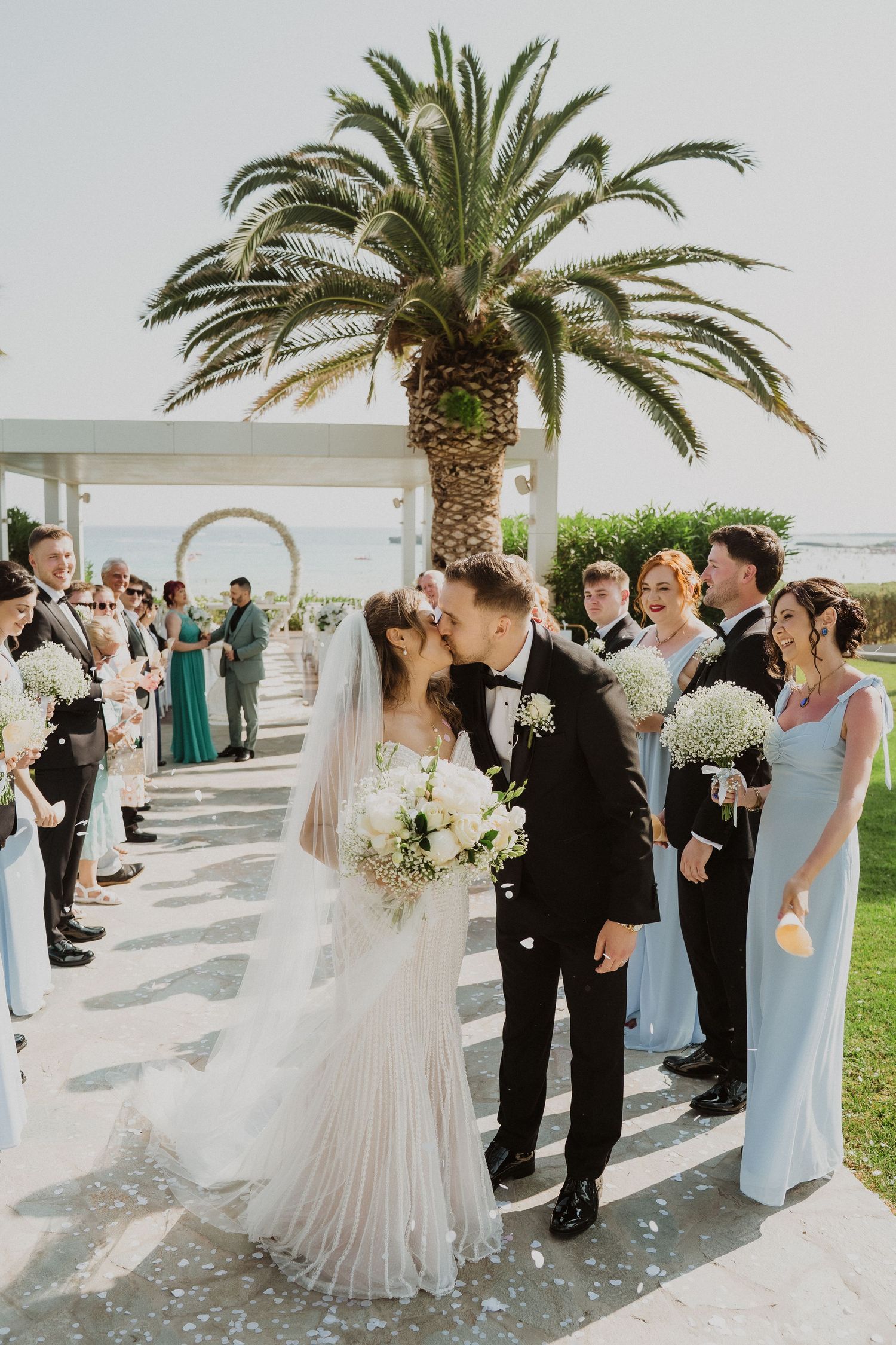 Newlyweds share a romantic kiss at outdoor wedding ceremony aisle lined with bridesmaids in blue and groomsmen.