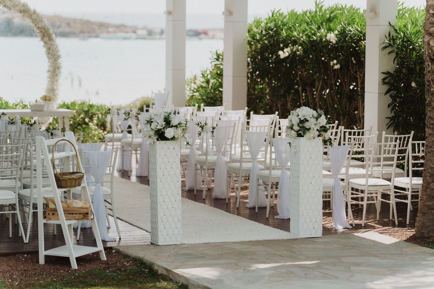 Elegant outdoor wedding ceremony setup with white chairs, floral arrangements, and a white aisle runner overlooking a scenic waterfront.
