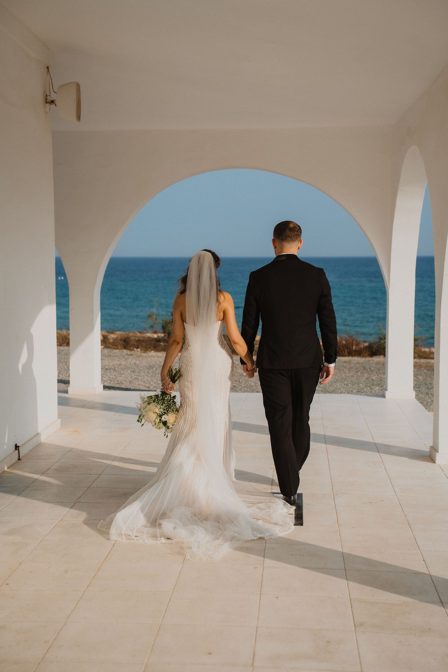 Bride and groom walking hand in hand through white arched colonnade toward stunning blue ocean at seaside wedding venue.