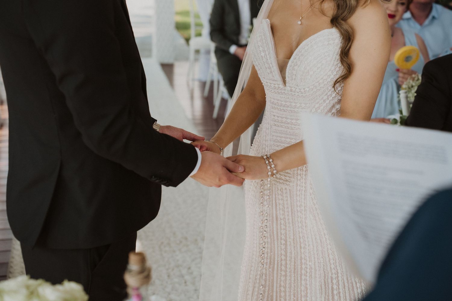 Close-up of a bride in a beaded white wedding dress holding hands with her groom during the ceremony.