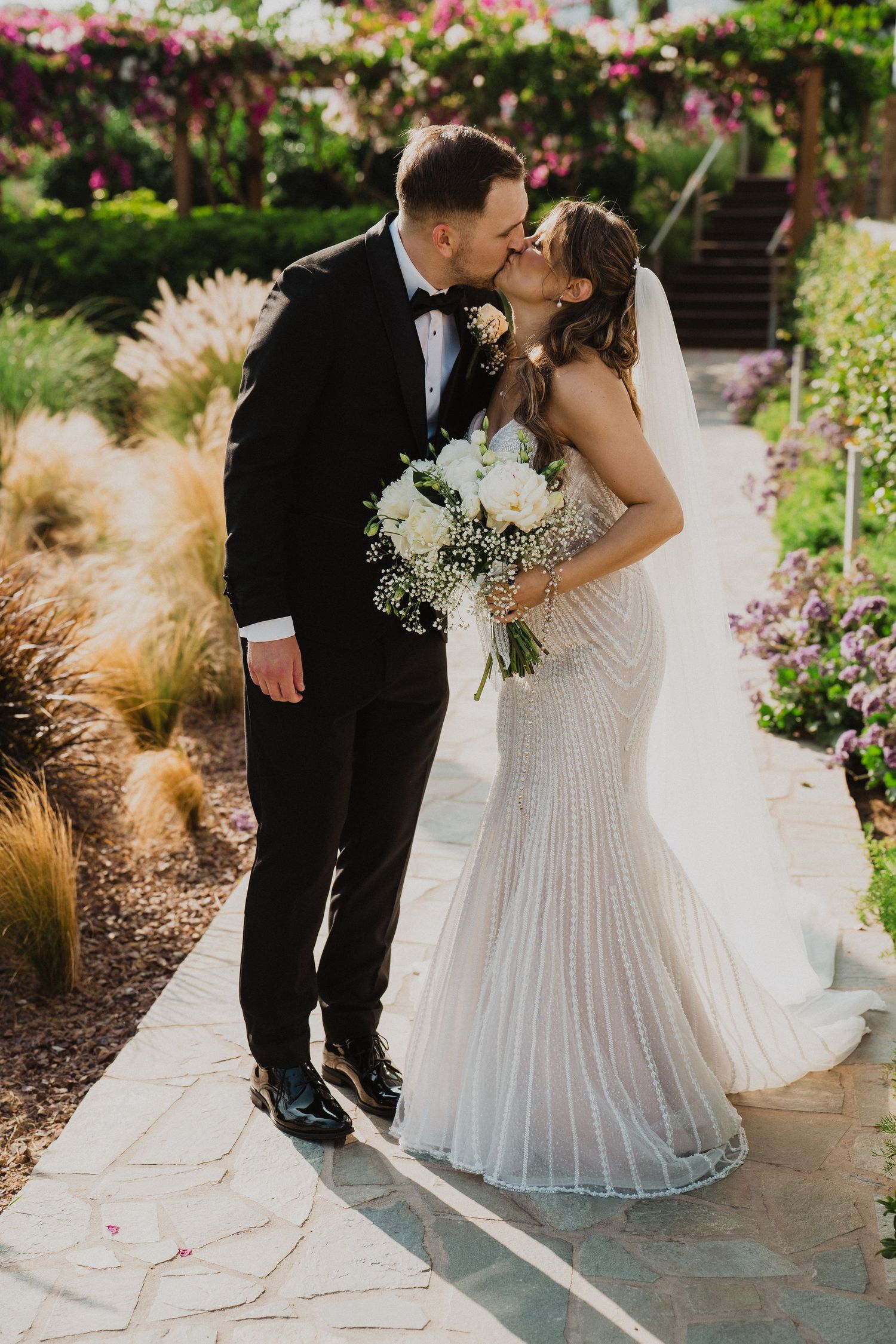 Newlyweds share romantic kiss on garden pathway surrounded by colorful flowers and lush greenery.