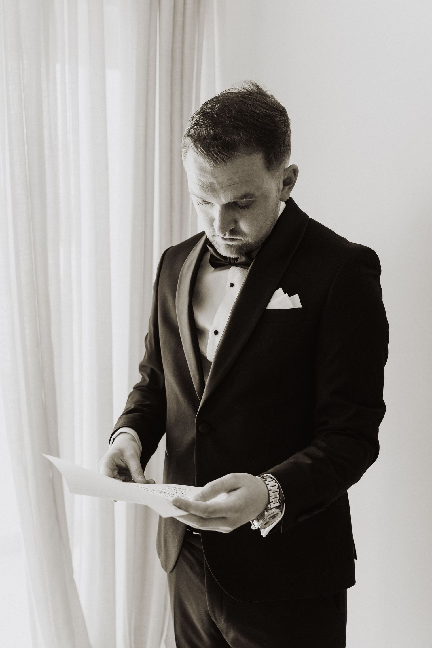 Groom in black tuxedo reading a heartfelt letter before wedding ceremony.