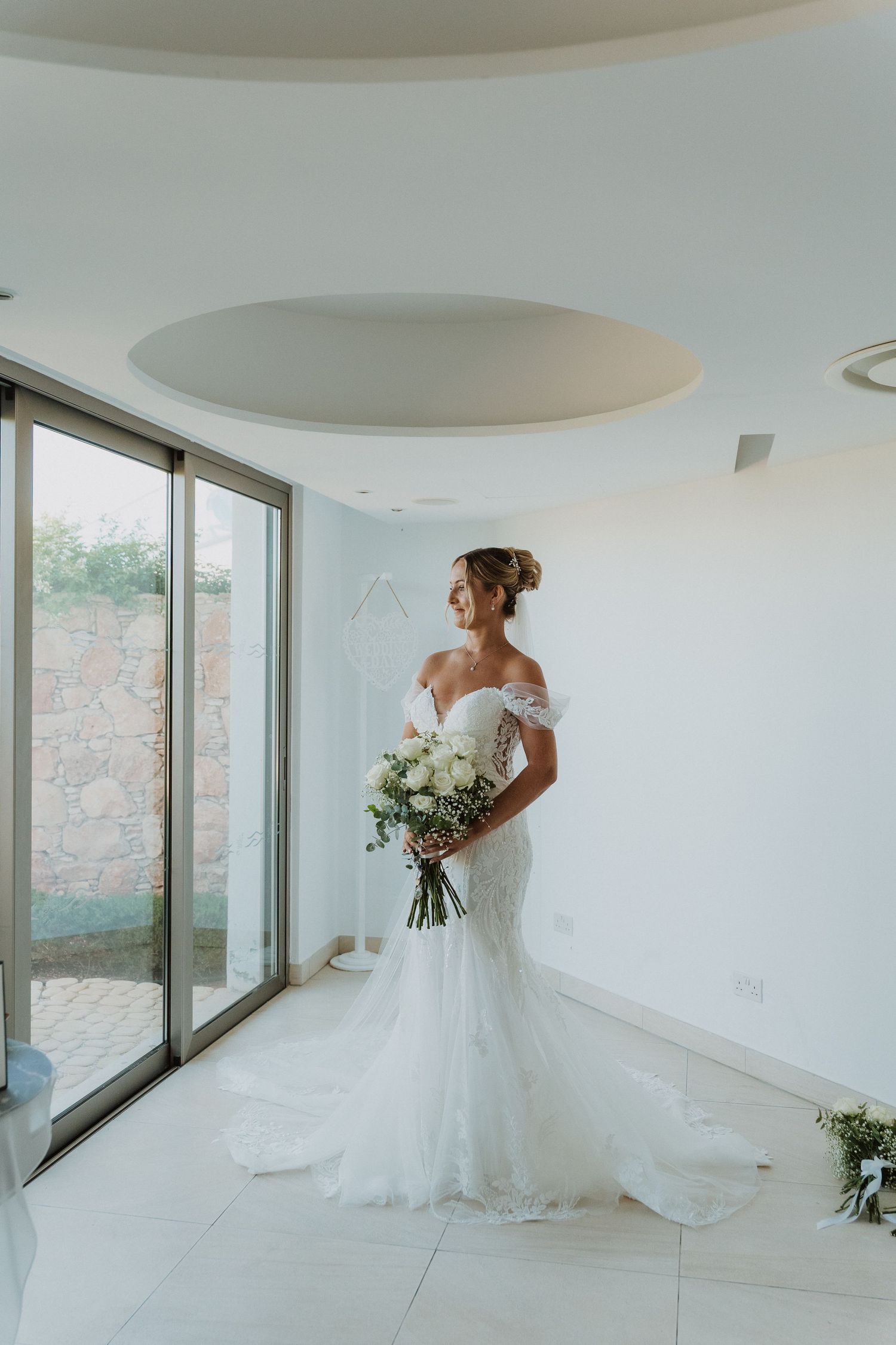 A bride in an elegant off-shoulder mermaid wedding gown holds a white bouquet while standing in a modern white room.