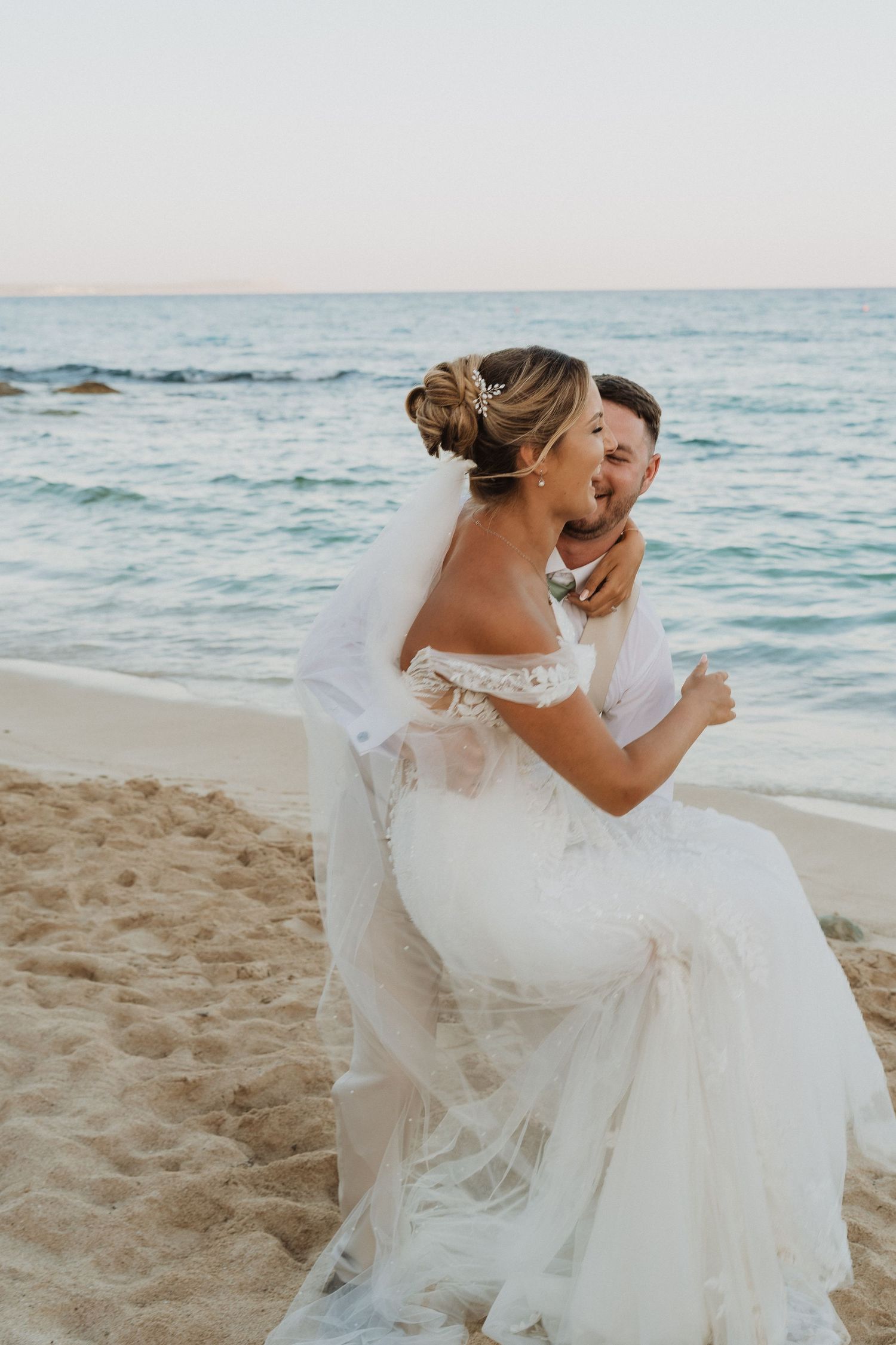 A bride in a flowing white lace dress and veil dances joyfully on a beach at sunset with ocean waves in the background.
