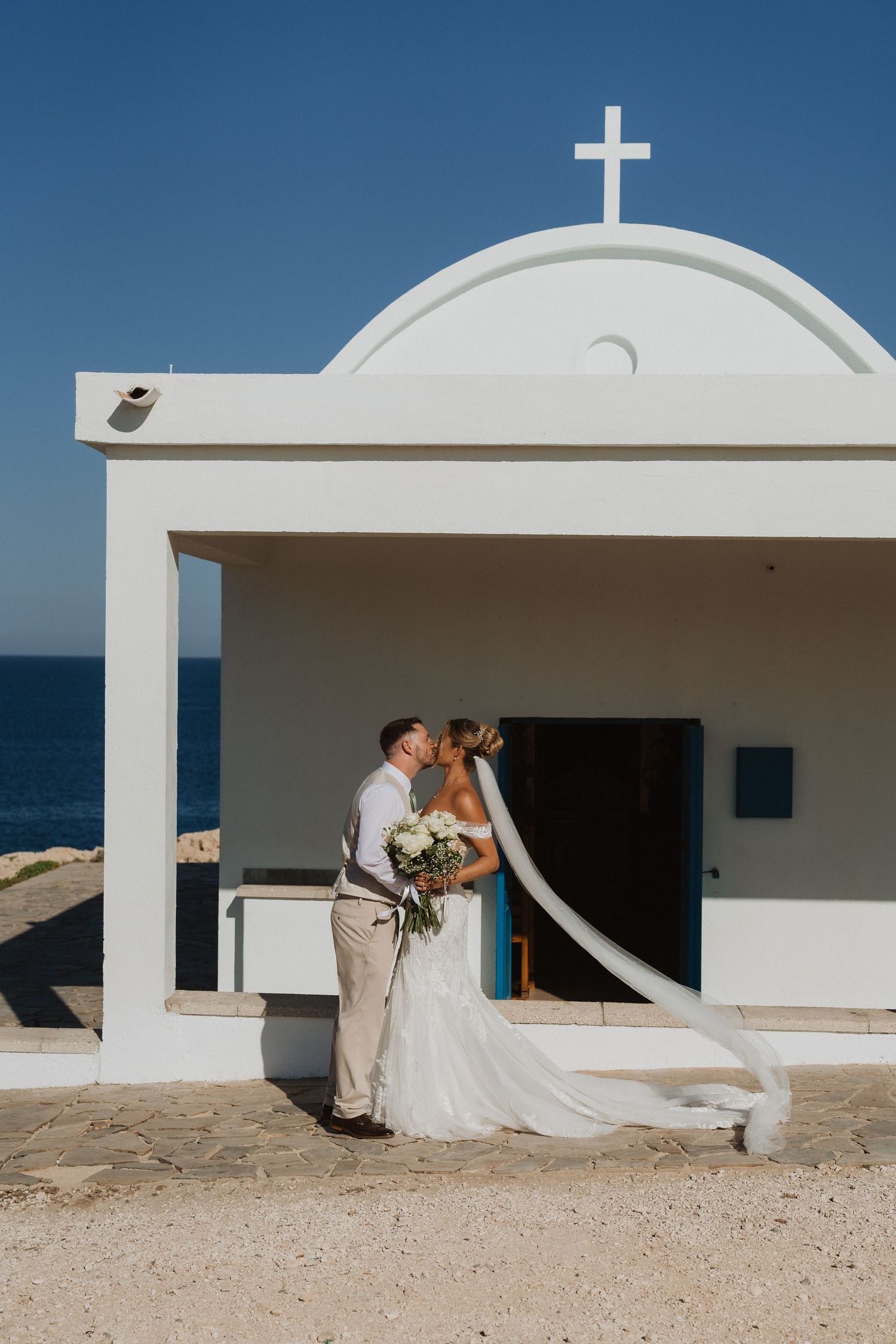 A romantic wedding couple embraces against white Mediterranean architecture and blue skies in a coastal setting.