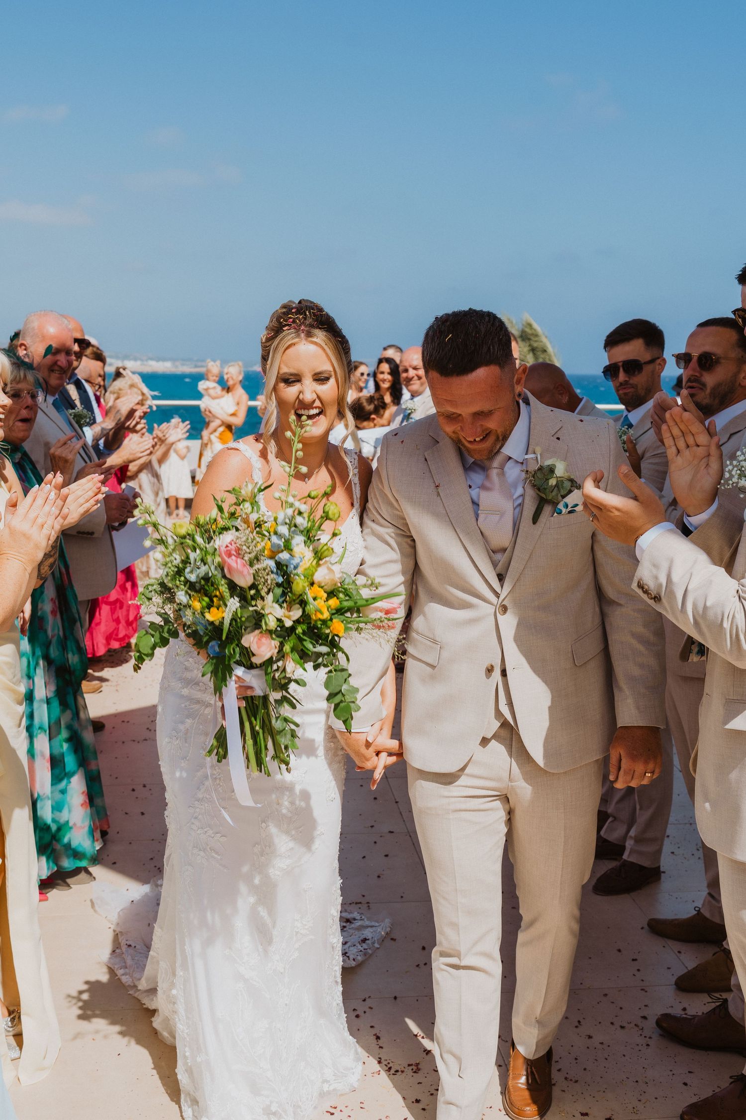 Newlywed couple walks down beach aisle while wedding guests cheer and throw confetti in tropical destination ceremony.