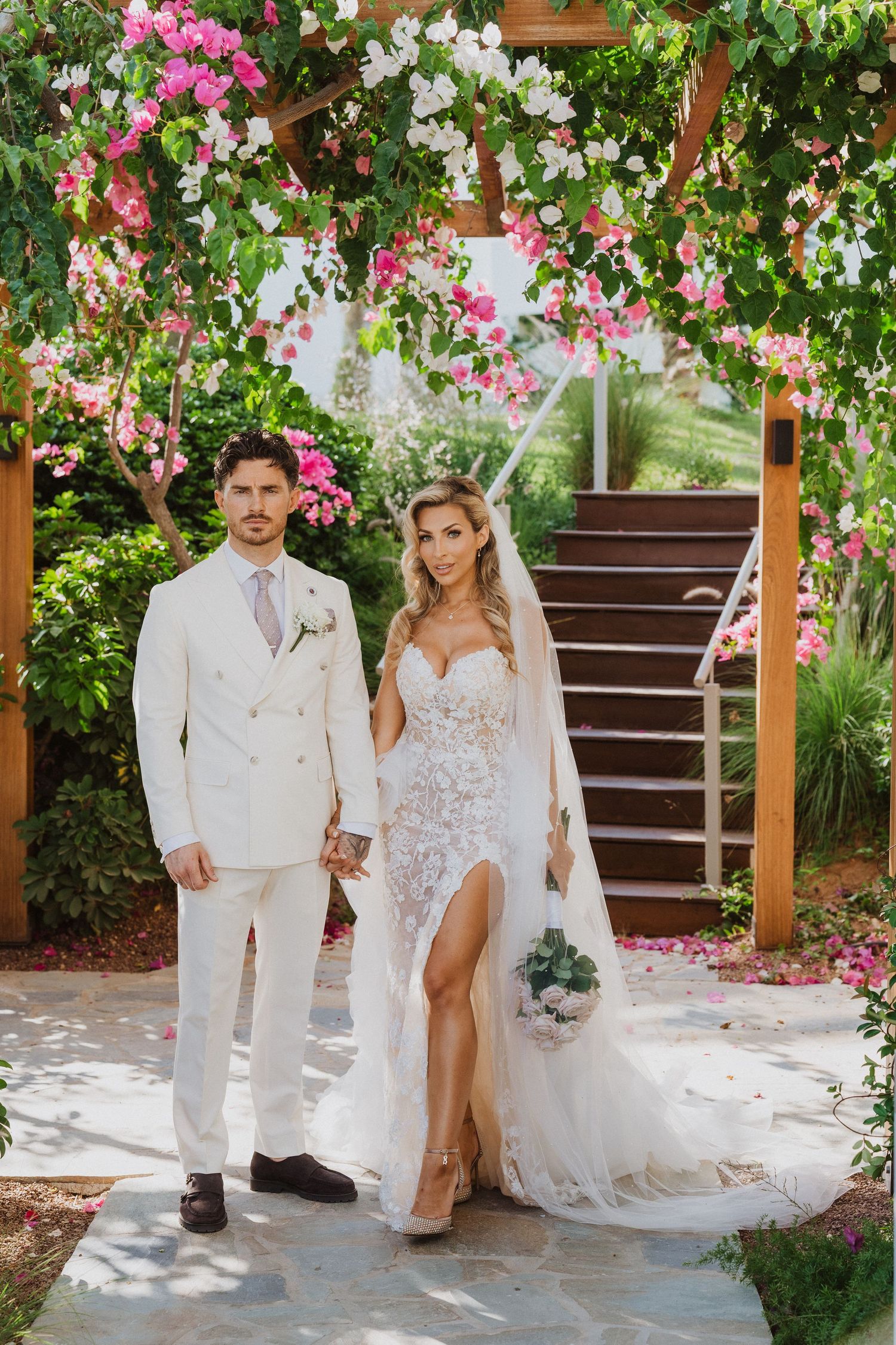 Wedding couple poses under beautiful pink floral arch in garden setting with white attire and romantic ambiance.