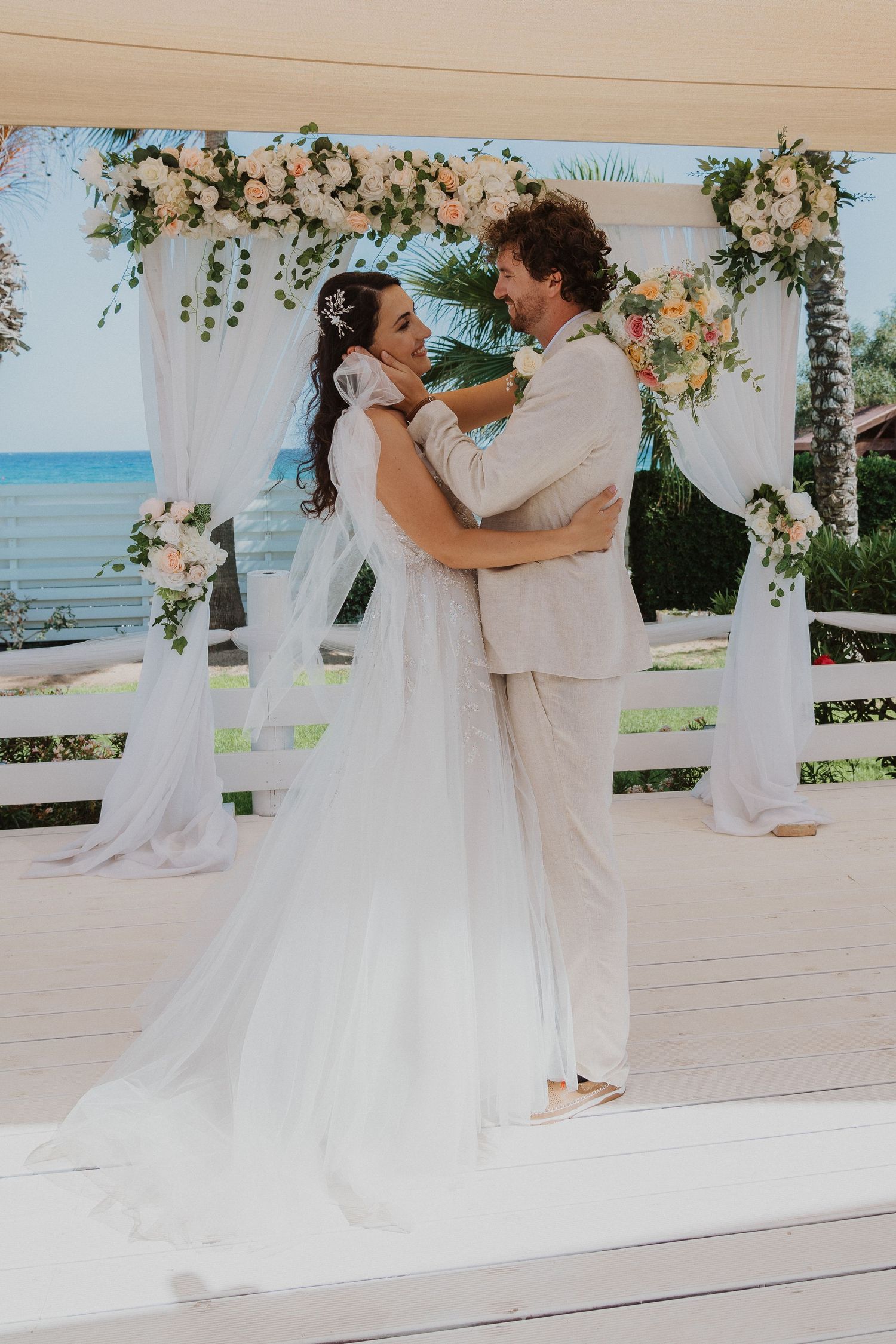 Dancing couple in wedding attire embrace under decorated arch with ocean in background.