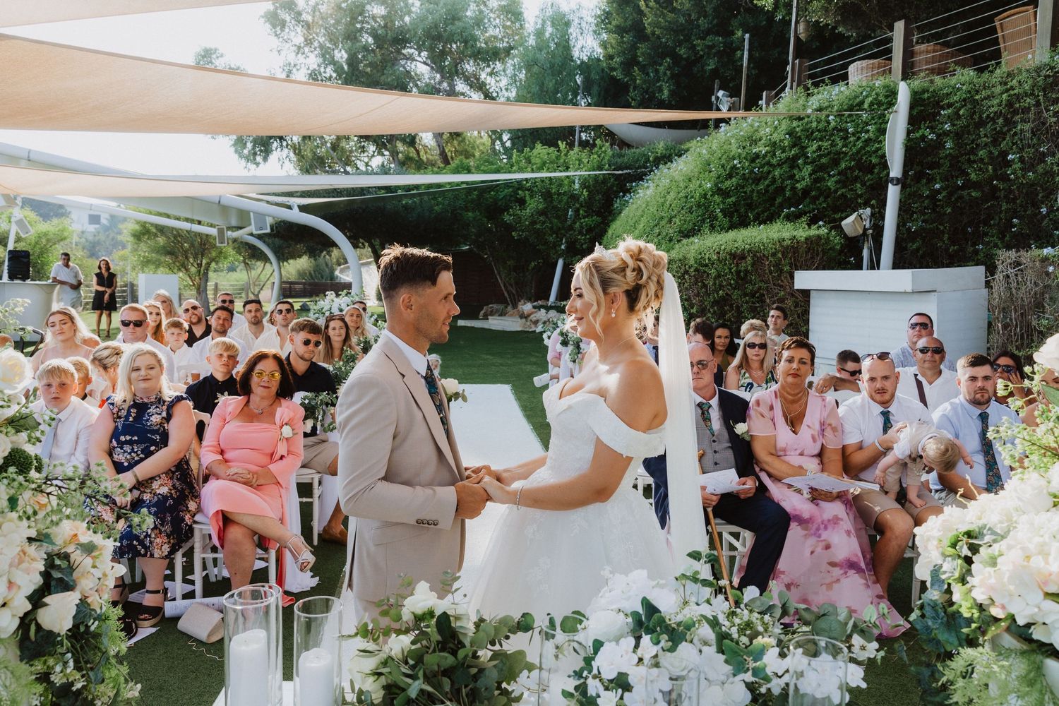 Wedding ceremony in a garden setting with guests seated and white floral decorations under shade sails.