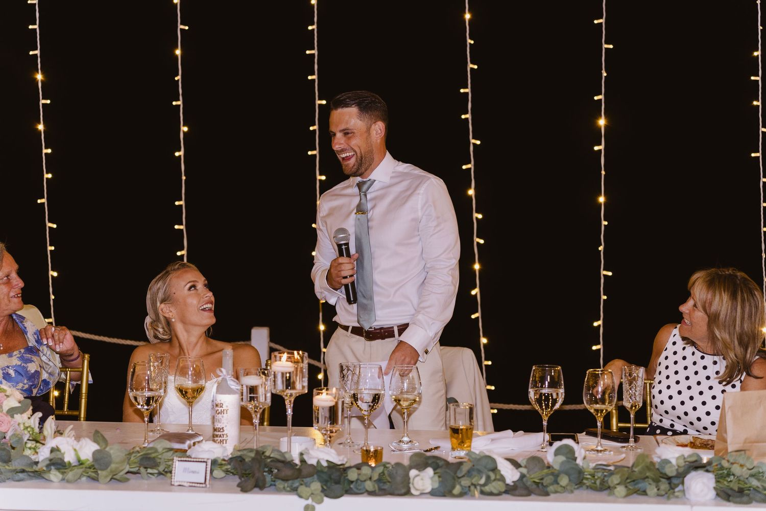 Wedding toast being delivered at head table decorated with greenery and lights.