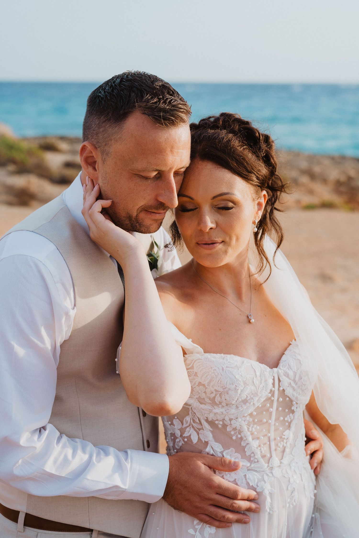 A romantic beachside wedding couple embraces in white attire against a turquoise ocean backdrop.